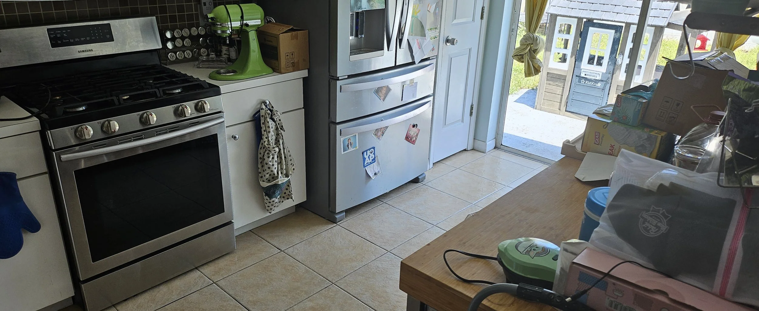 A messy kitchen with a stainless steel stove, a white counter with a green mixer, a gray refrigerator with magnets, and a sliding glass door leading outside. Various food packages and kitchen items are cluttered on the right side.