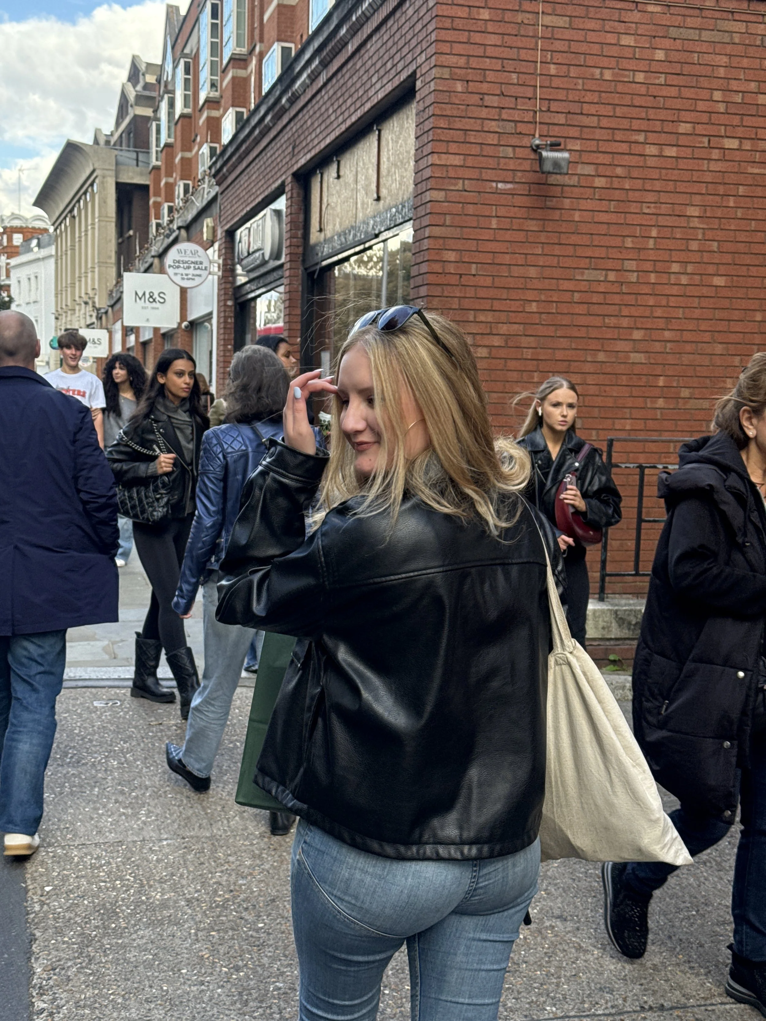 A woman with blonde hair, wearing a black leather jacket and blue jeans, is in focus, smiling and adjusting her sunglasses while walking on a busy London city street with other pedestrians, brick buildings, and shop signs in the background.