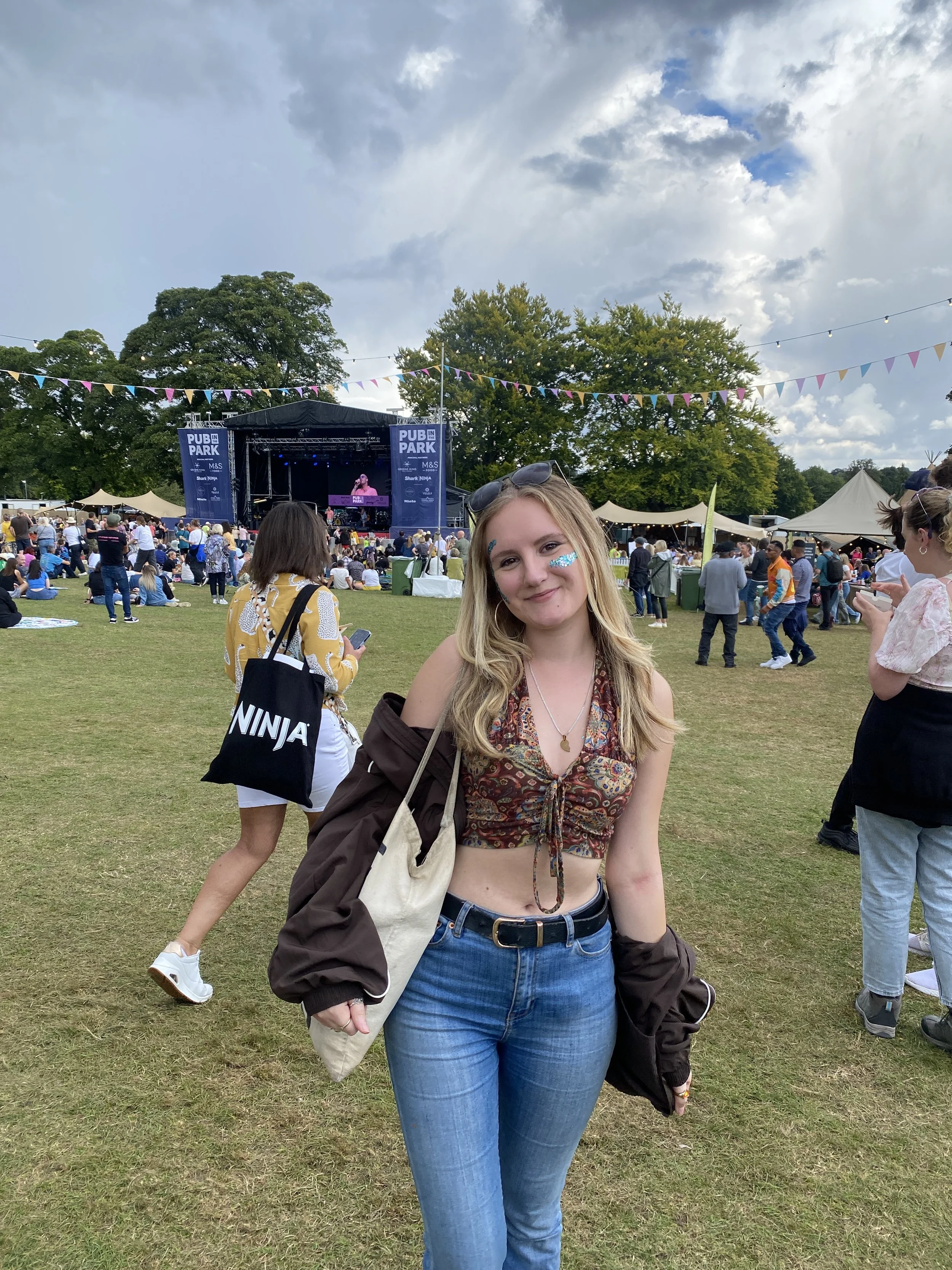 Young woman with face glitter at Pub in the Park, wearing a floral crop top, blue jeans, and sunglasses on her head, standing at an outdoor music festival with a stage and crowd in the background.