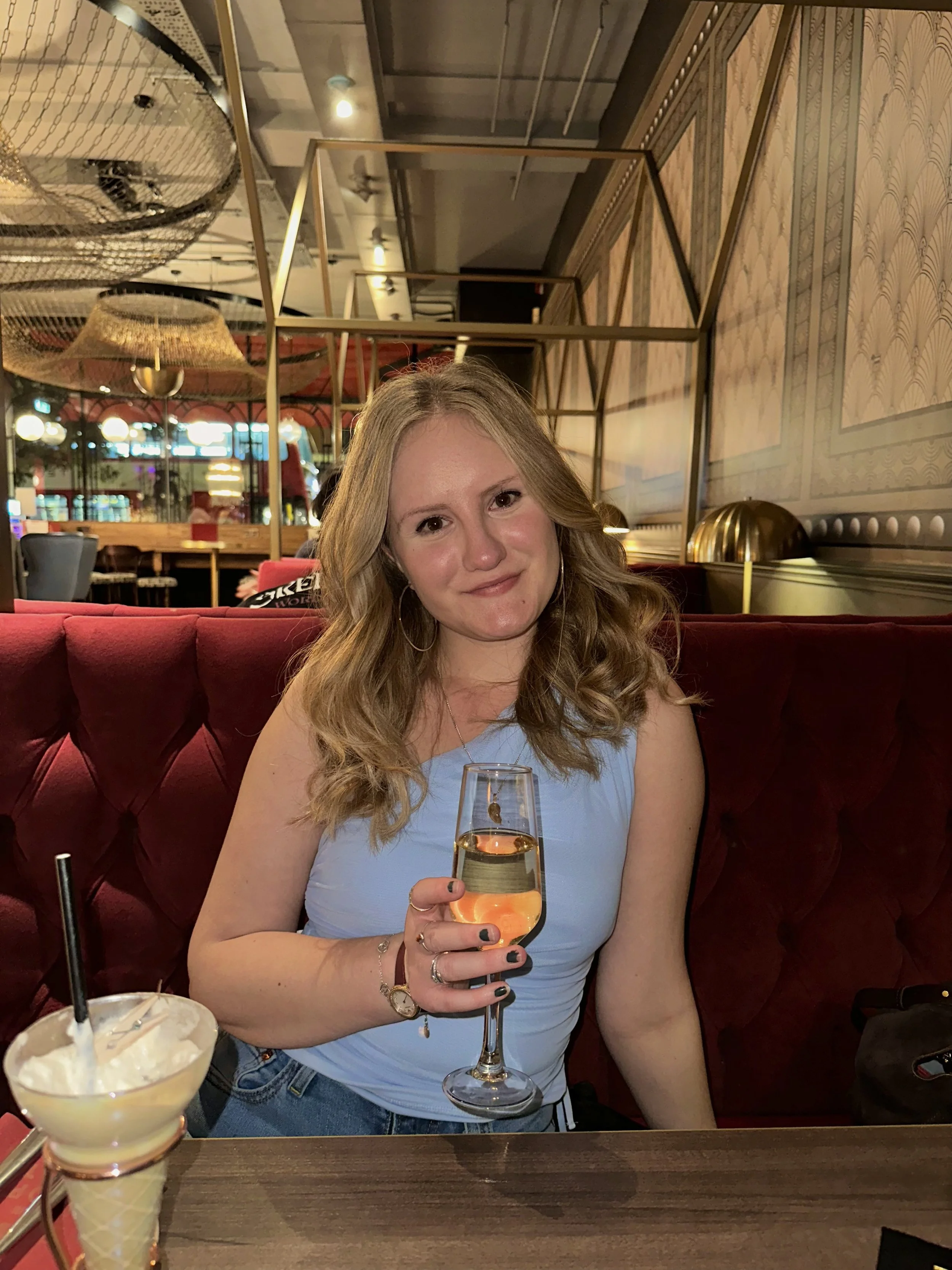 A woman with long wavy blonde hair, wearing a light blue sleeveless top, sitting on a red tufted booth in a restaurant or bar in London, specifically The Top Hat Bar, holding a glass of champagne.