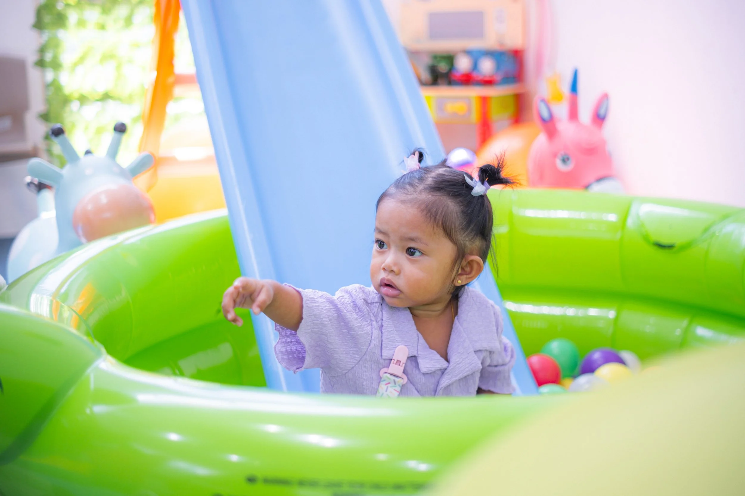 Young girl sitting in a green inflatable pool with a blue slide, pointing to something outside the frame, with colorful balls around her in a playroom decorated with inflatable animal figures.