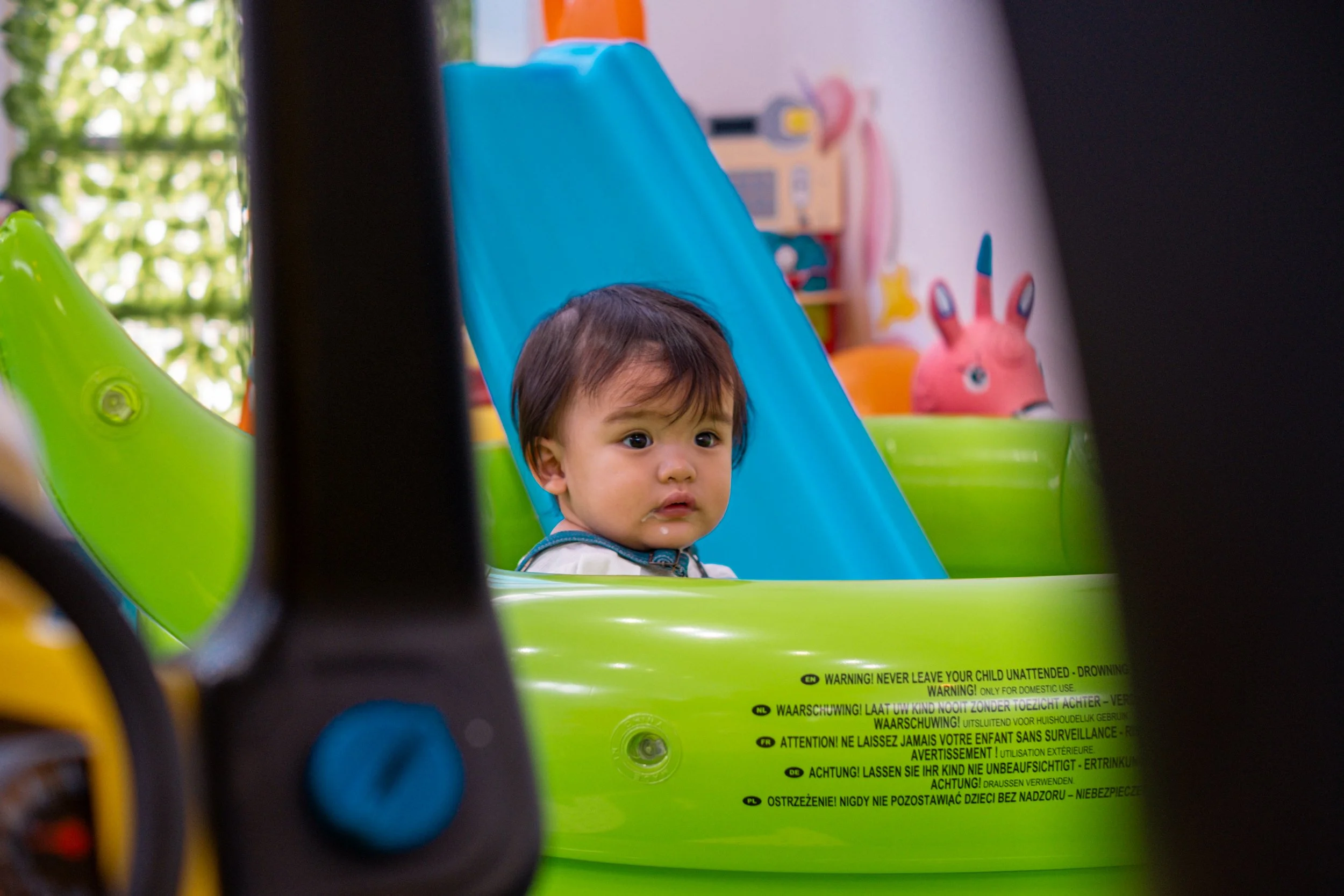 A young child with dark hair sitting in a green and blue toy stroller, looking to the side with a curious expression. In the background, there is a pink plush toy in the shape of a rabbit and colorful decorations in a playroom.