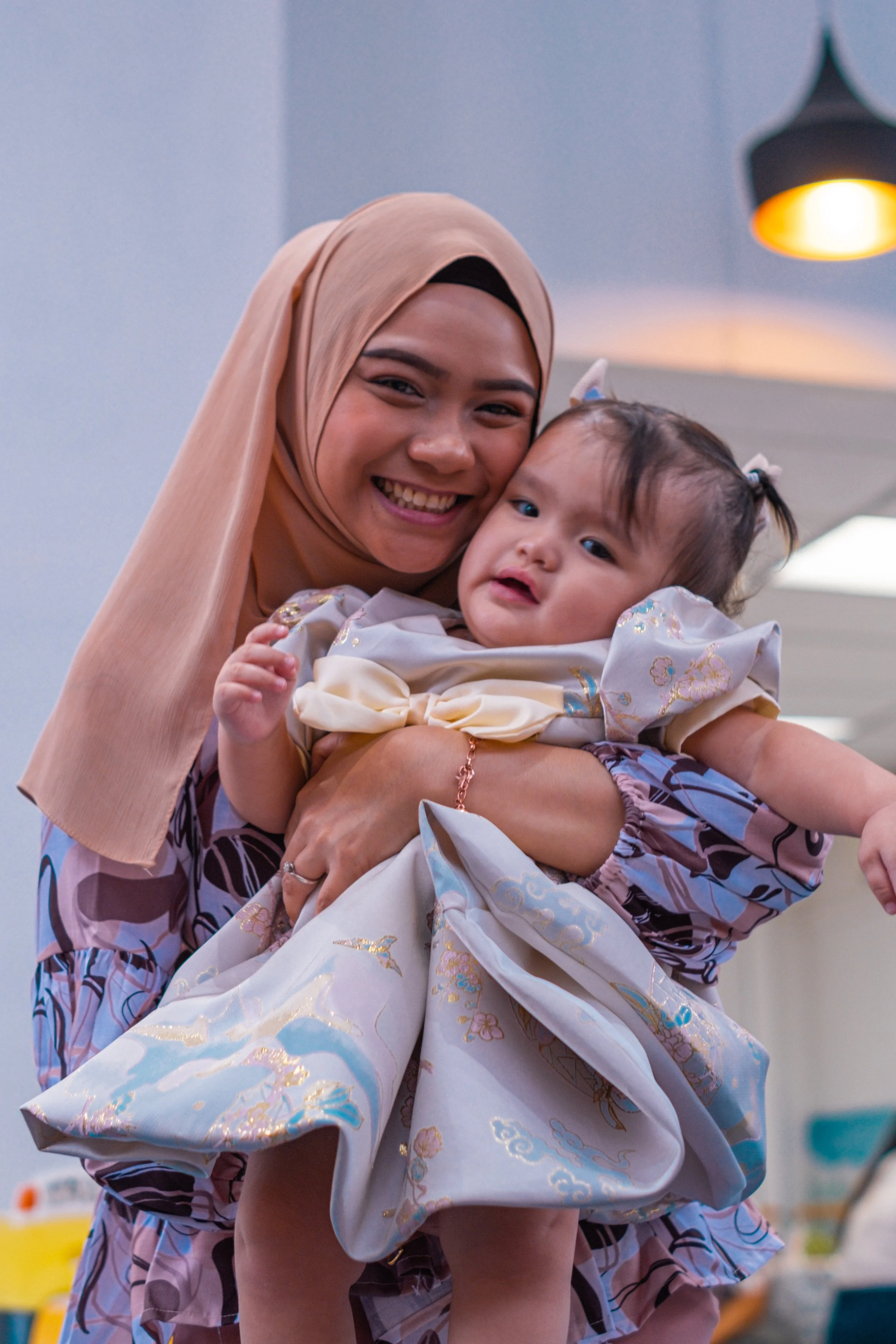 A woman holding a young girl, both smiling and looking at the camera, in an indoor setting with warm lighting.