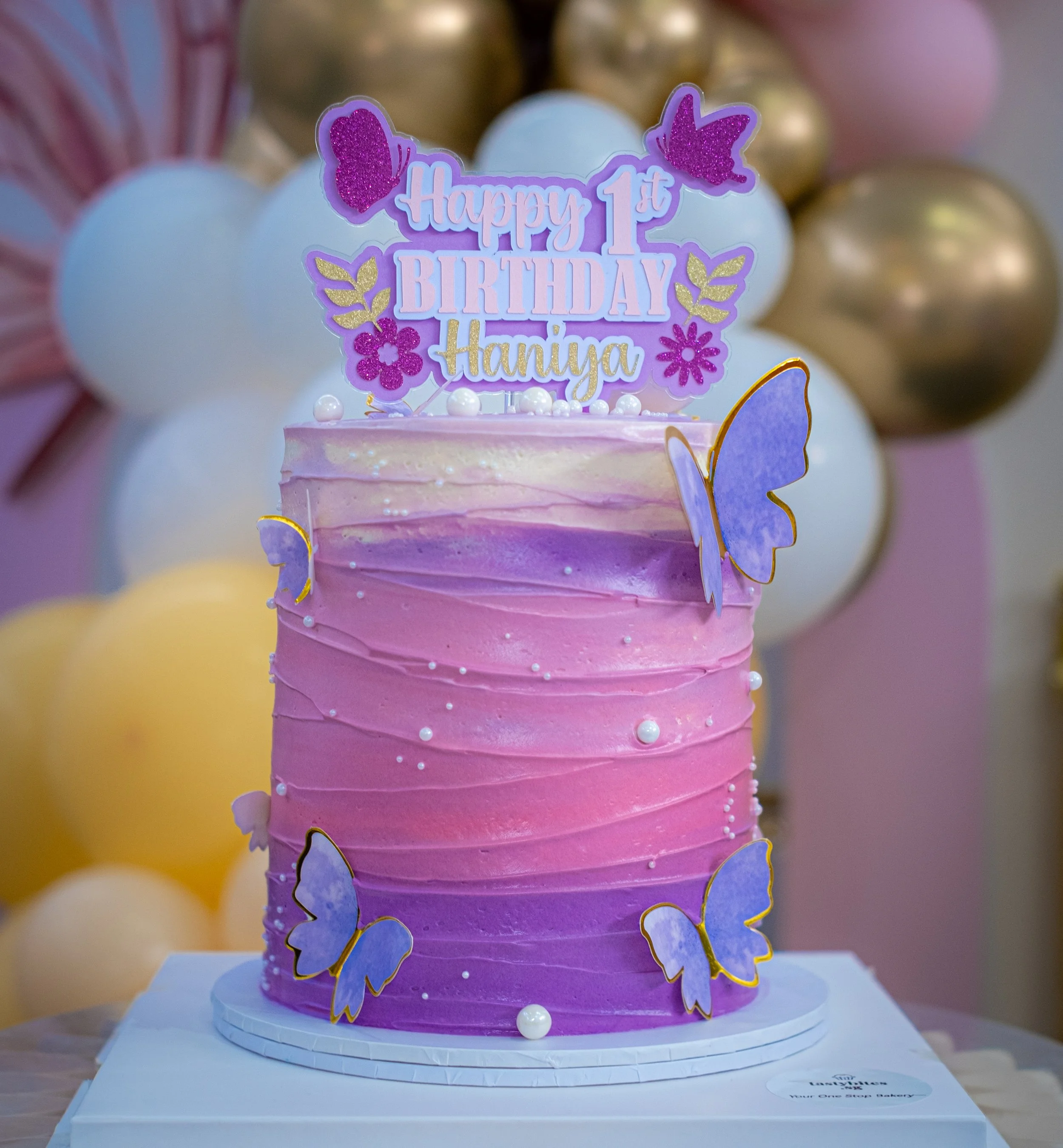 A pink and purple ombré birthday cake decorated with purple butterflies and white pearls, with a topper reading 'Happy 1st Birthday Haniya', surrounded by pastel balloons.