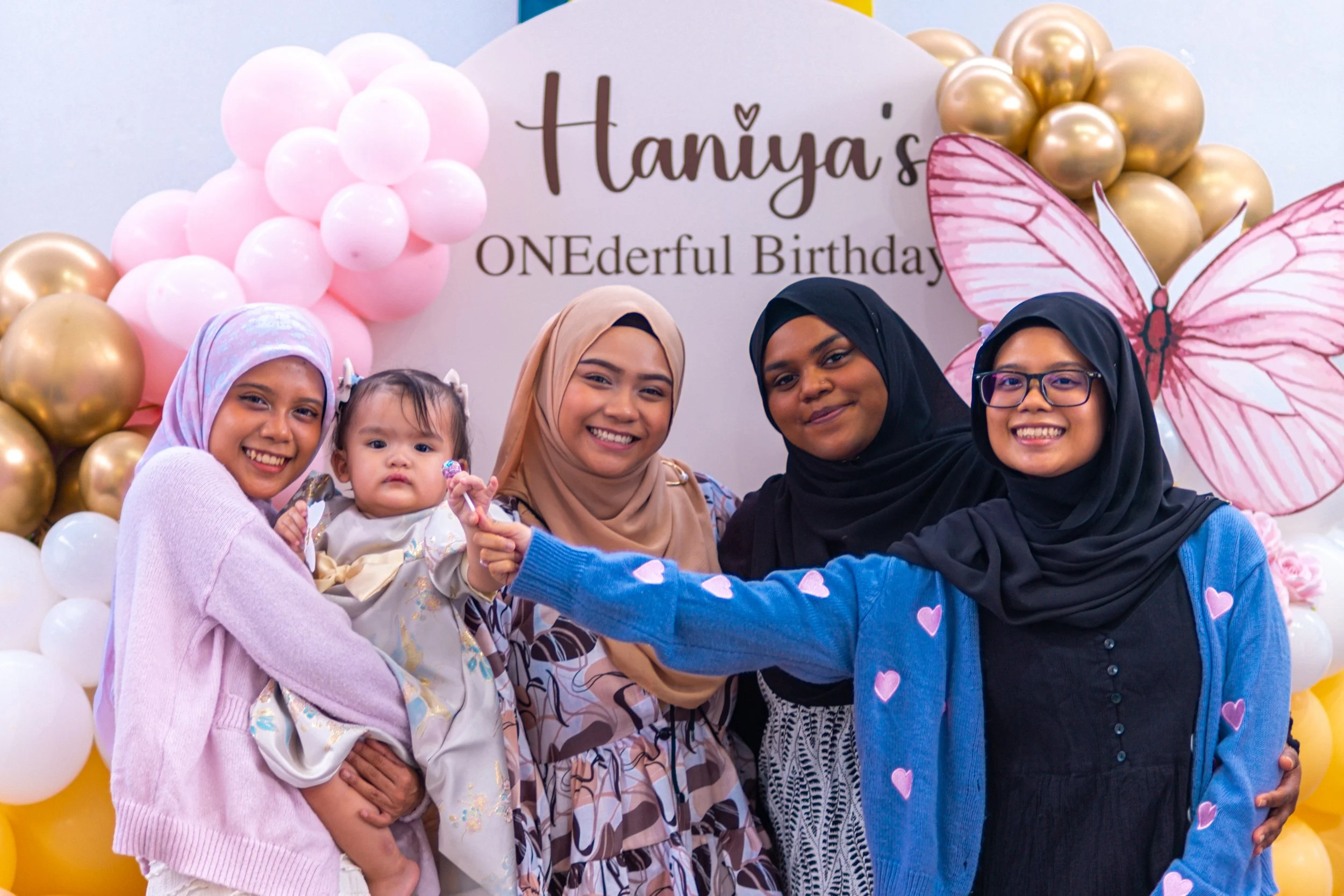 A group of five women and children smiling at a birthday celebration with pink and gold balloons, a butterfly cutout, and a sign that reads 'Haniya's One-derful Birthday'.