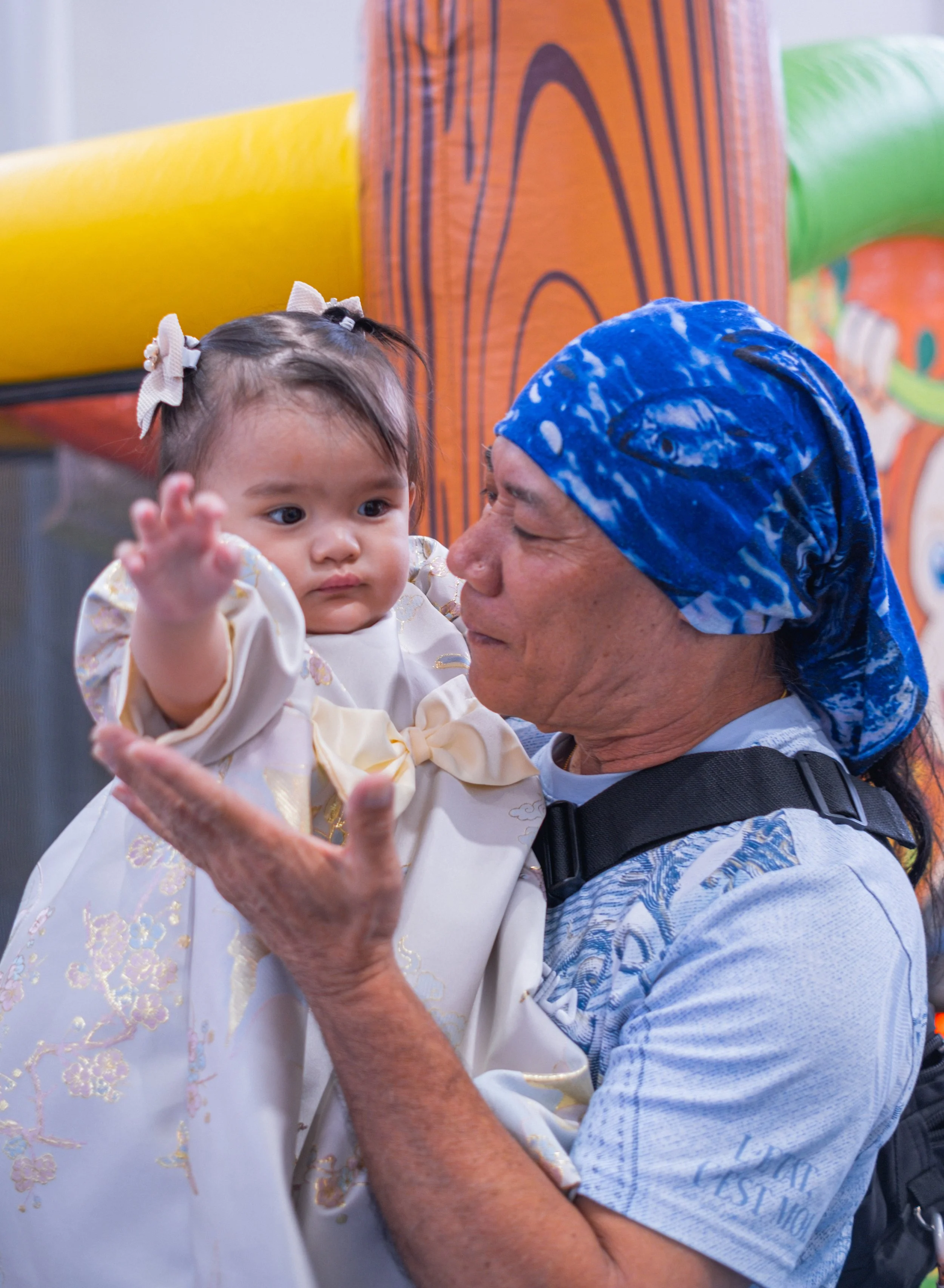 An older man with a blue headscarf holding a young girl in a white dress with floral embroidery, both indoors against a background with colorful play structures.