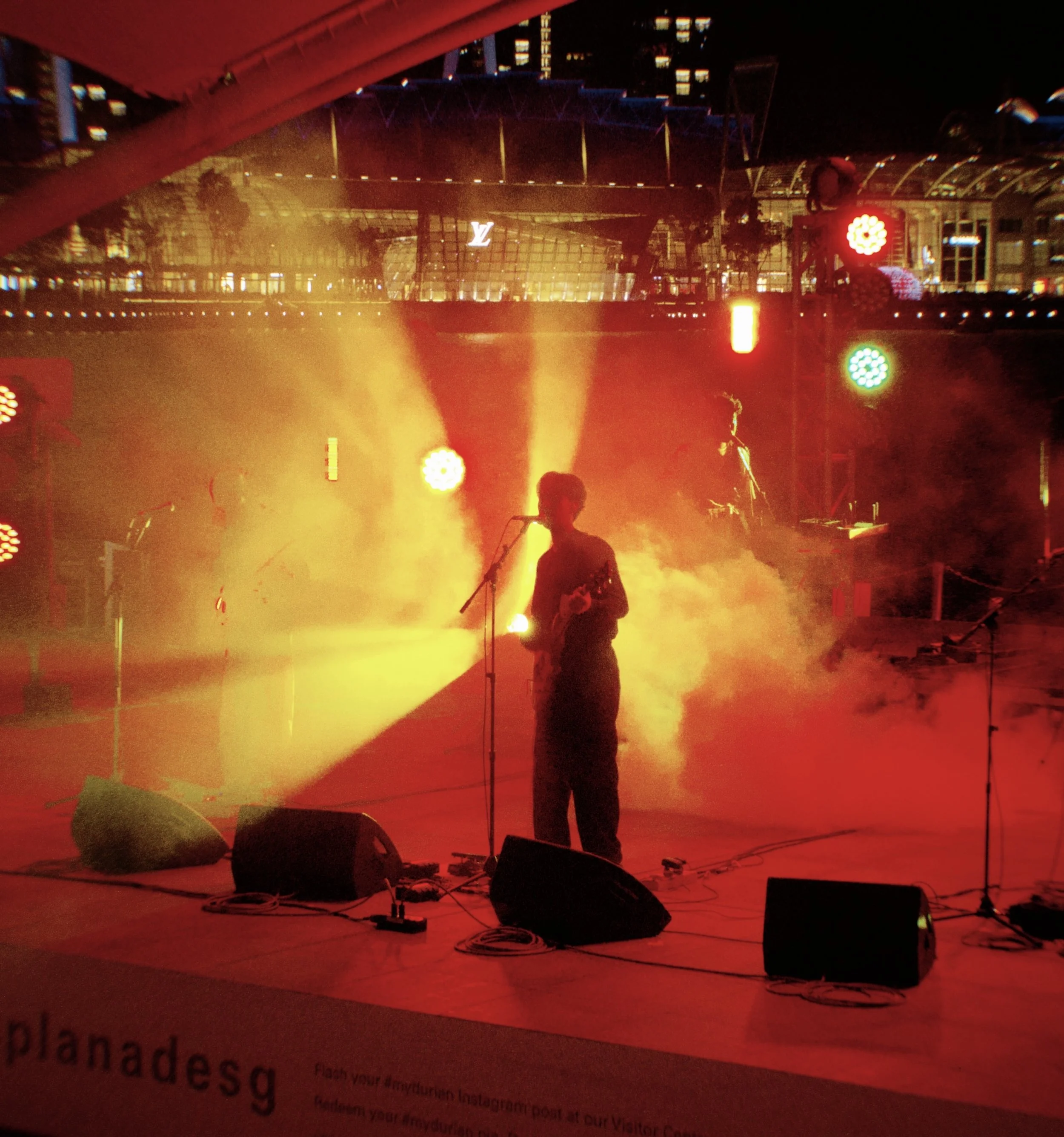 A musical performance on a stage with colorful lights and fog, featuring a silhouette of a person playing a guitar and singing into a microphone.