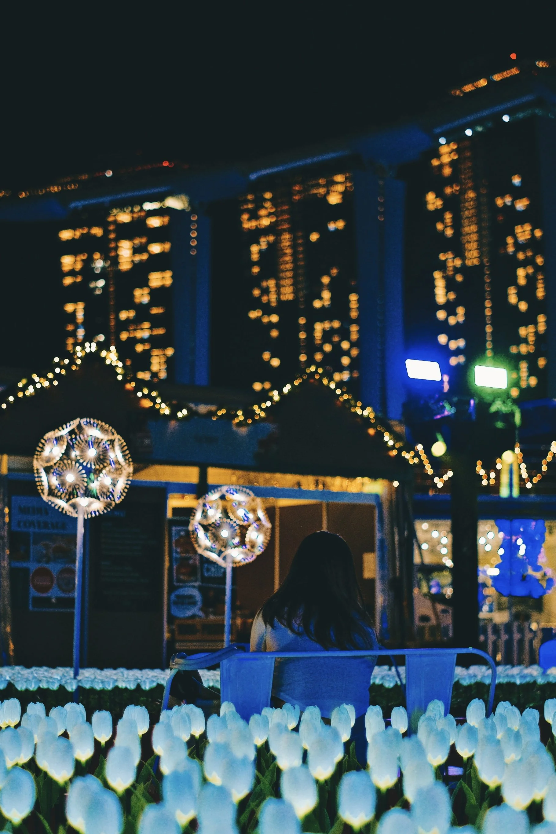 A woman sits on a bench in a flower bed of white tulips at night, with decorated illuminated spherical lanterns and string lights around her, and a high-rise building with lit windows in the background.