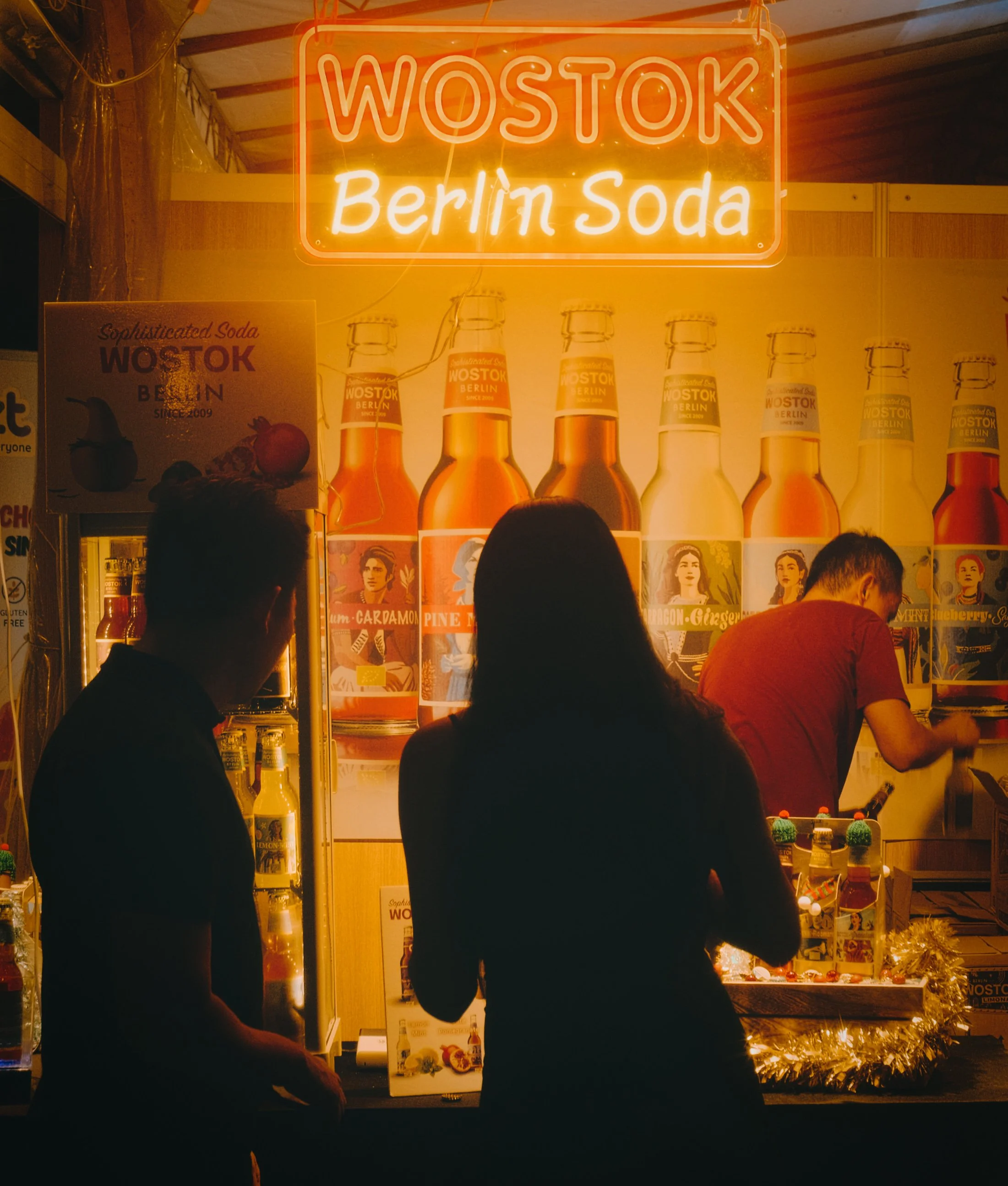 Silhouettes of two people in front of a brightly lit stand selling bottled beverages, with neon signs stating 'WOSTOK' and 'Berlin Soda' above.