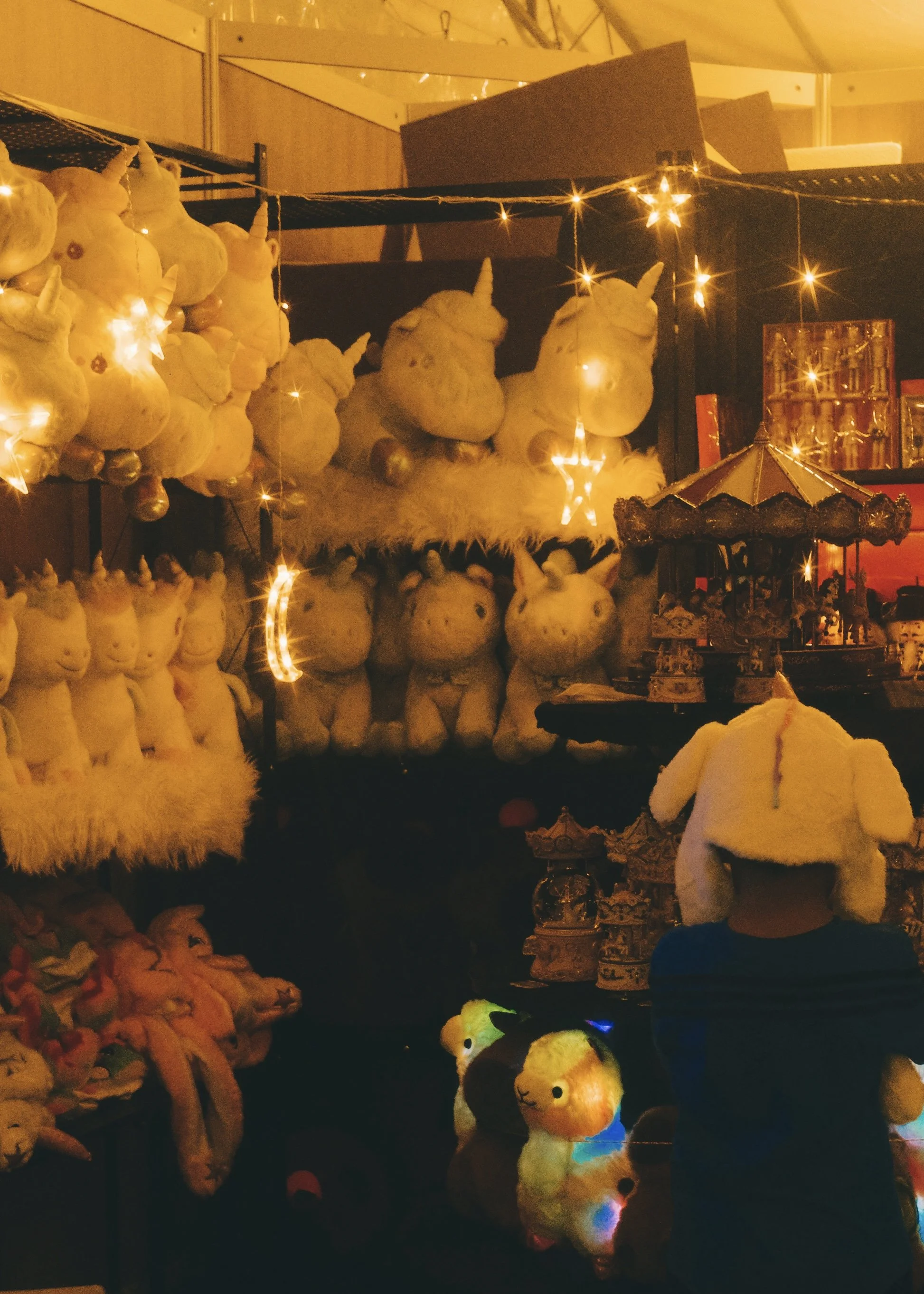 A display of plush unicorn toys on shelves, decorated with fairy lights at a market stall, with a child wearing a unicorn hat in the foreground.