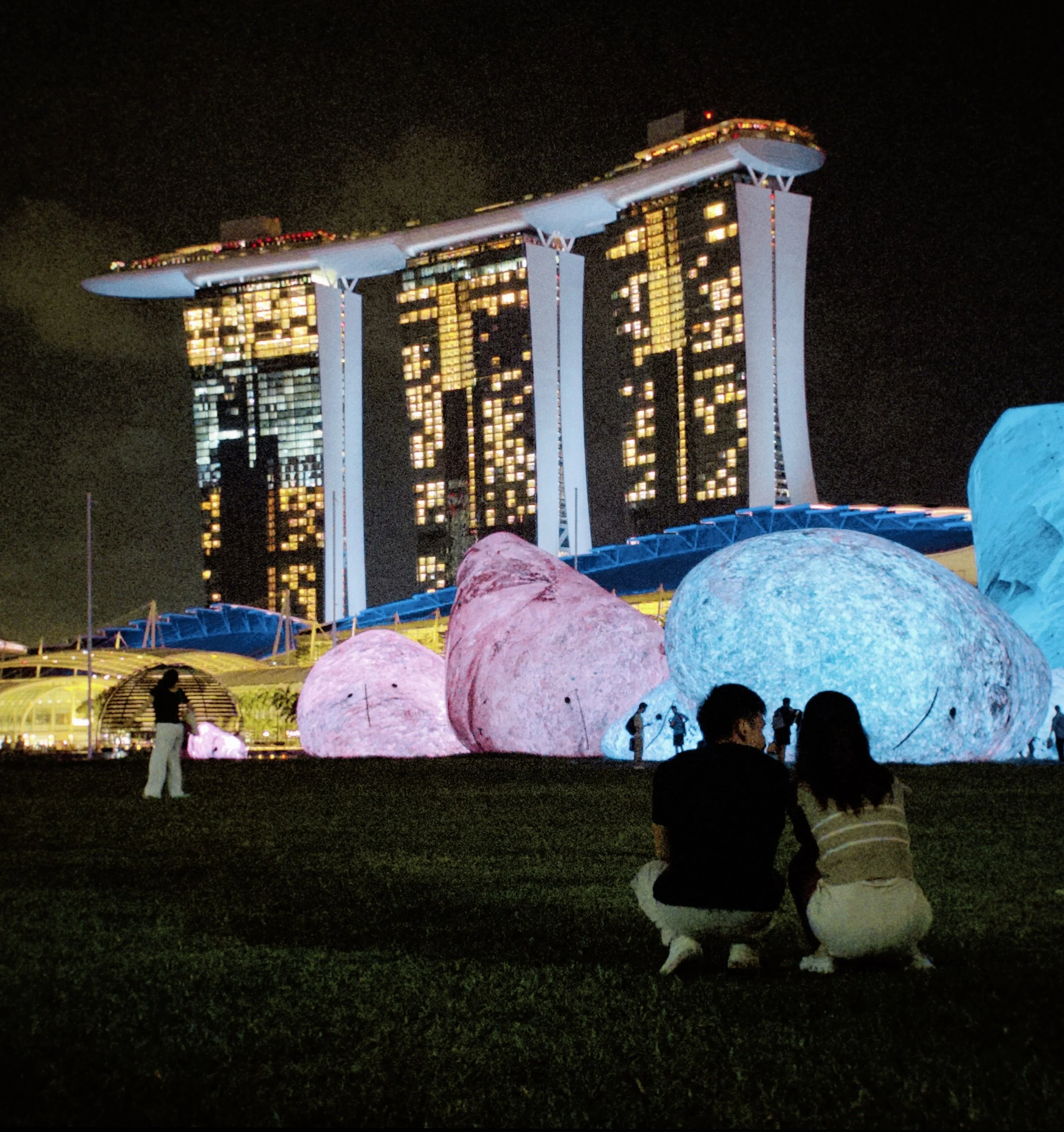 Night view of the Marina Bay Sands hotel in Singapore with illuminated three tower structure, large decorative rocks in the foreground, and people sitting on the grass.