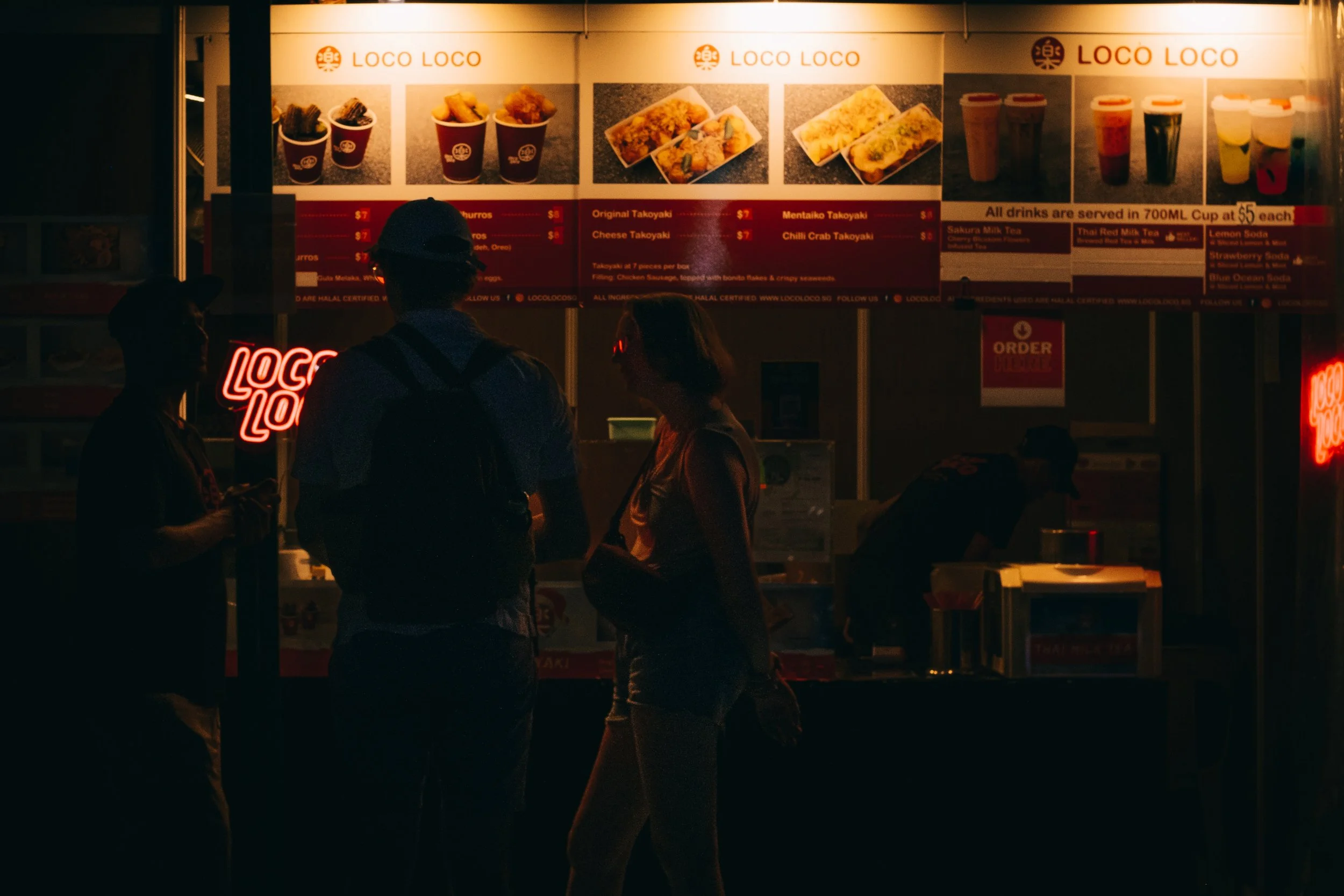 People ordering food at a brightly lit food stand called Loco Loco, with menu items and drinks displayed on signs above.