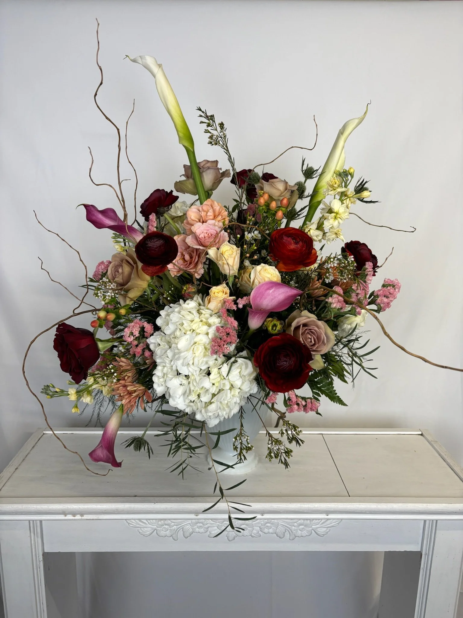 A colorful flower arrangement with roses, calla lilies, roses, orchids, and other flowers in a white vase on a white table against a neutral background.