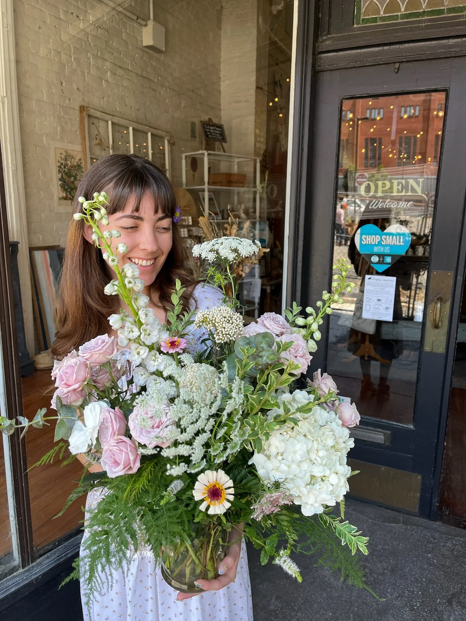Woman smiling and holding a large bouquet of mixed flowers outside a shop with a glass door that has an 'OPEN' sign and a 'Shop Small' sticker.