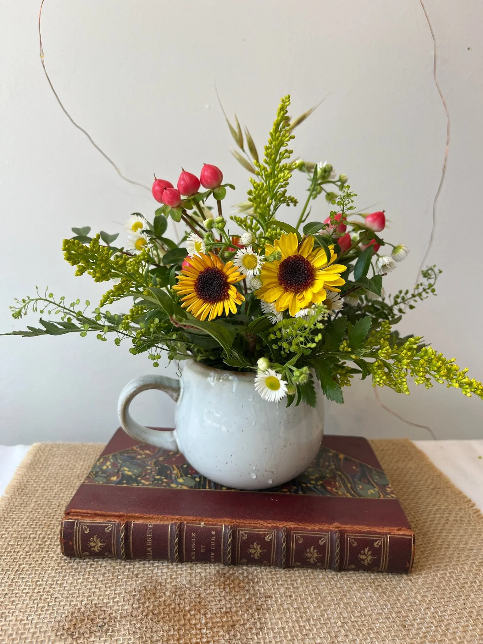 A bouquet of mixed flowers in a white ceramic mug, placed on top of a large, vintage red leather-bound book. The bouquet includes yellow daisies, small white daisies, pink berries, and green foliage. The arrangement is set on a burlap cloth with a wh