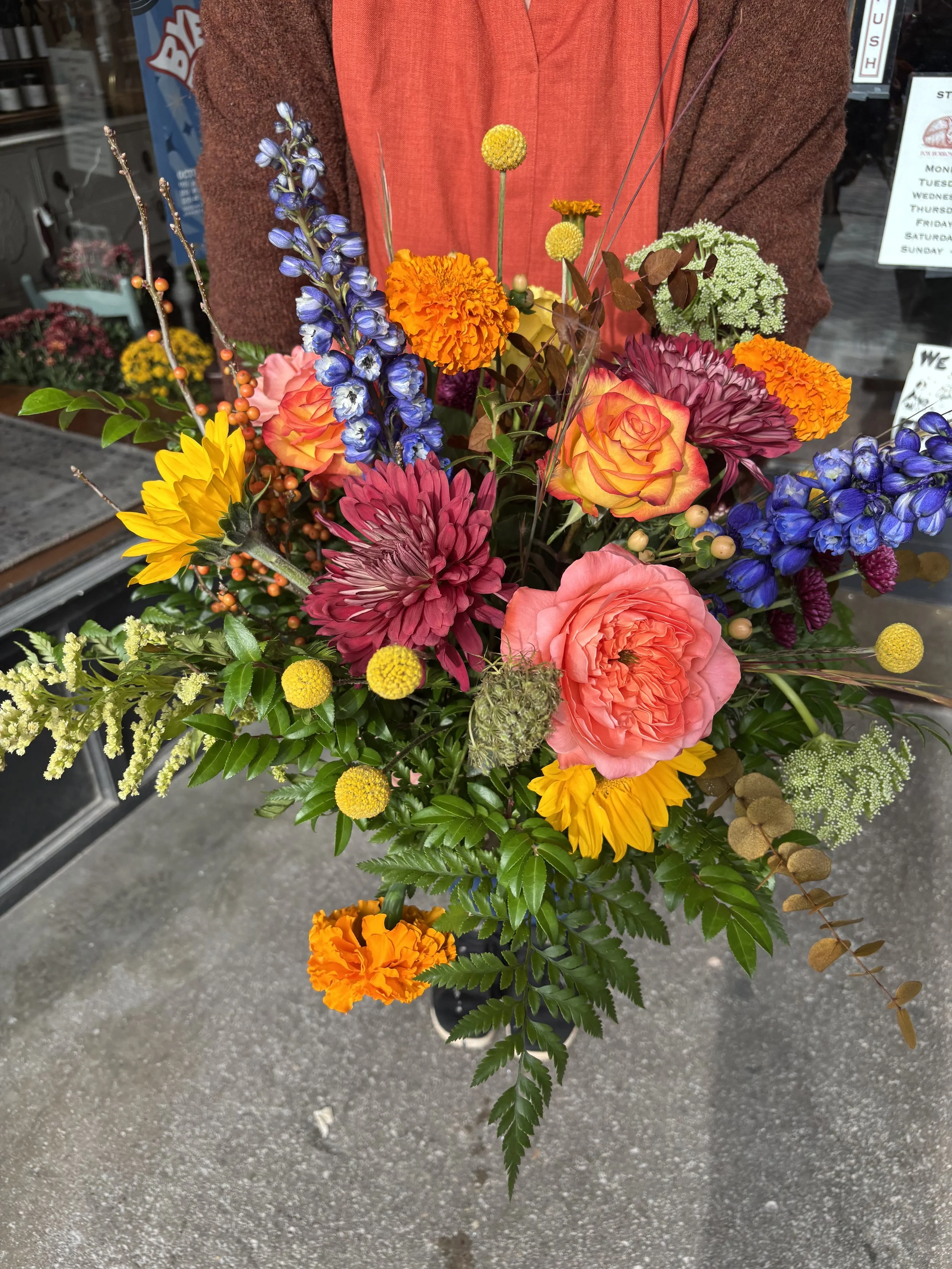 Vivid flower bouquet with roses, dahlias, marigolds, delphiniums, sunflowers, and various greenery carried by a person wearing an orange shirt and brown sweater outside a shop.