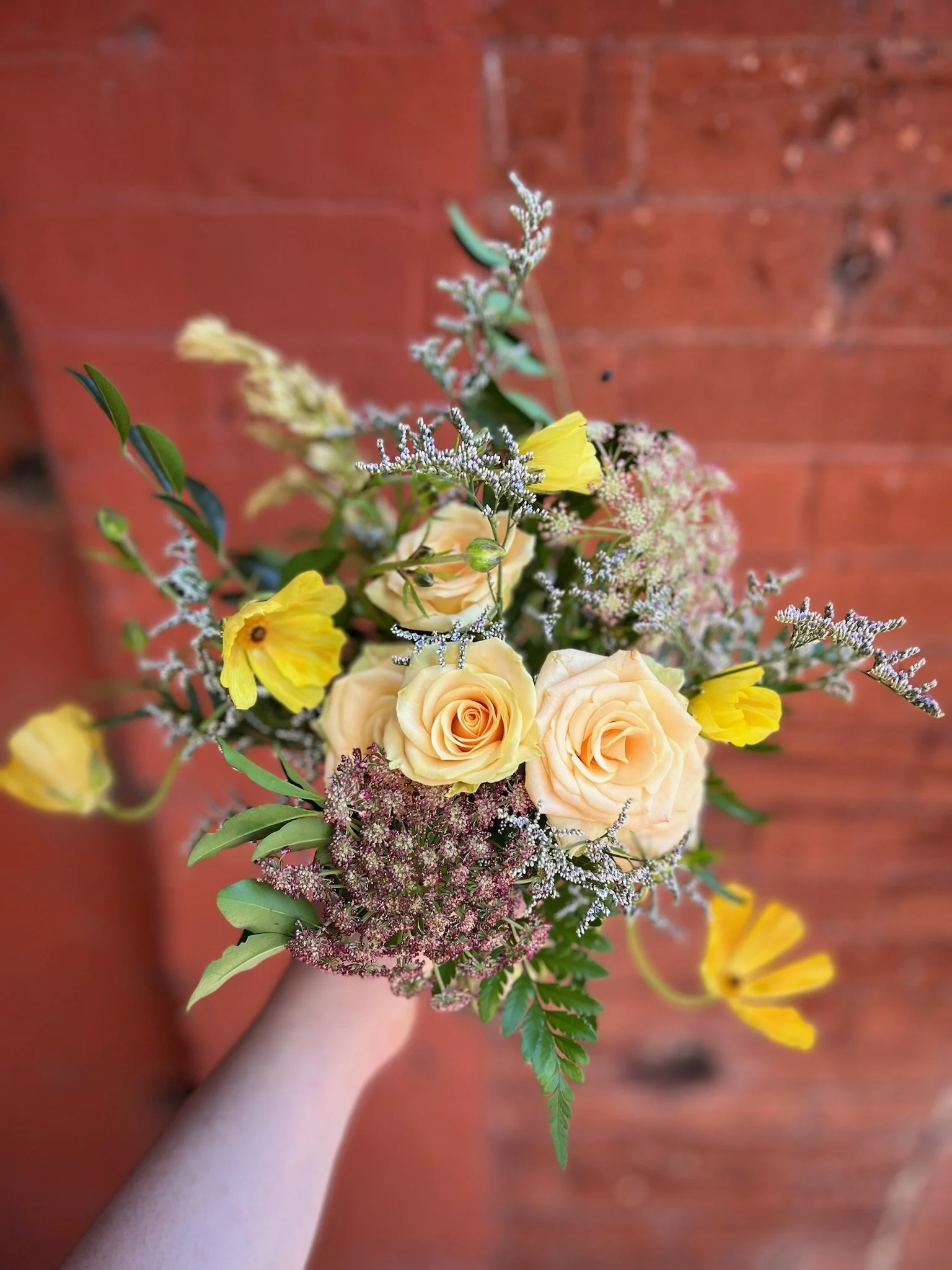 A hand holding a colorful bouquet of peach roses, yellow flowers, and greenery against a red brick wall background.