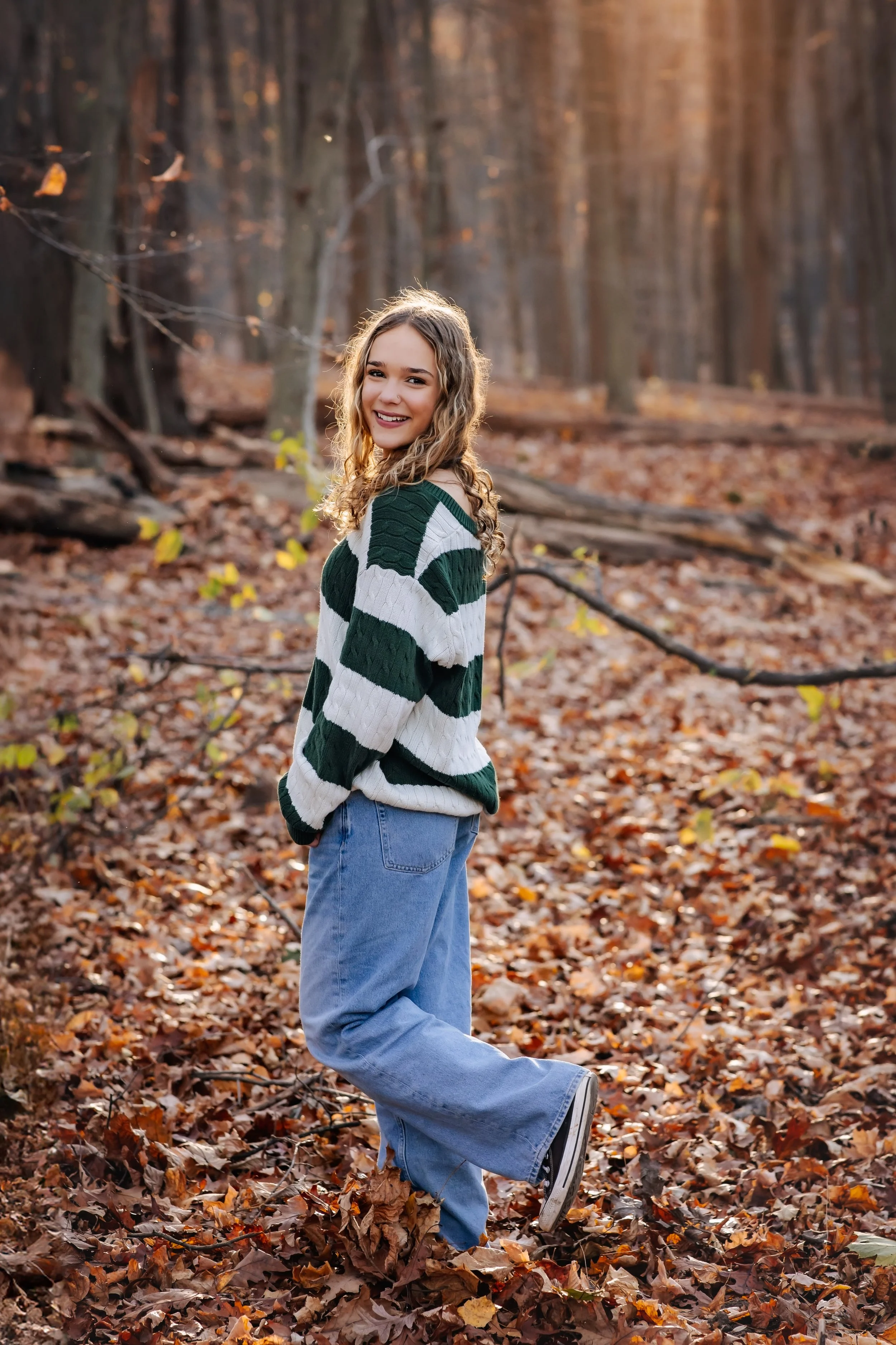 A young woman with curly light brown hair wearing a green and white striped sweater and blue jeans stands in a forest with fallen autumn leaves, smiling at the camera.