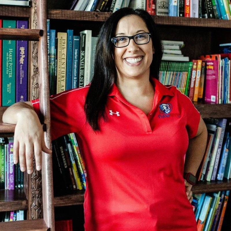 A woman with glasses and long dark hair smiling, wearing a red sports shirt, standing in front of a bookshelf filled with colorful books.