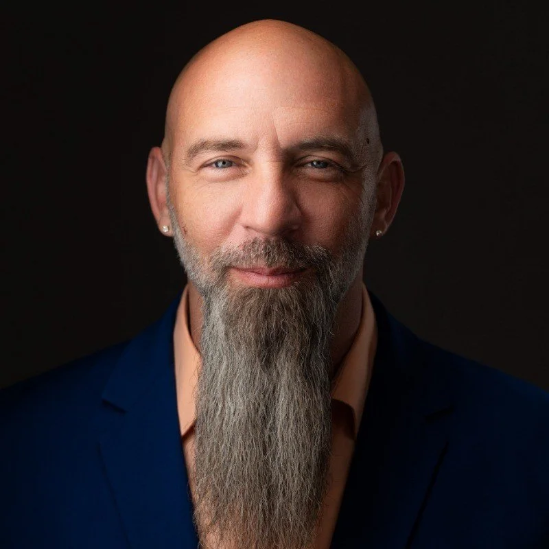 A portrait of a bald man with a long gray beard, wearing a dark blazer and a peach-colored shirt, against a black background.