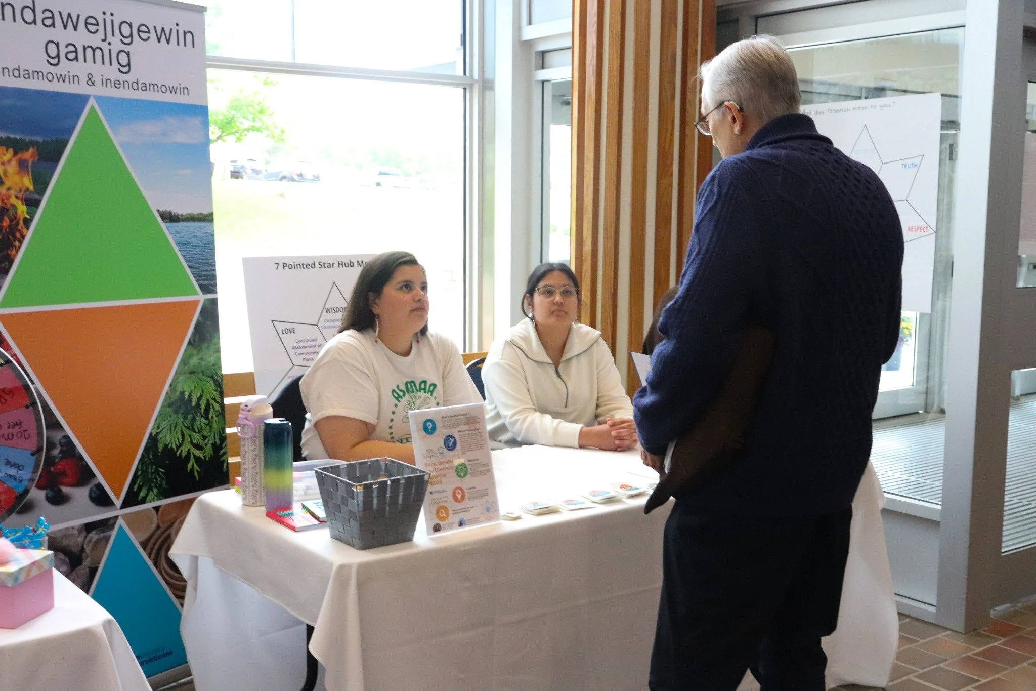 Two young women sitting at a table with informational materials, talking to an older man standing in front of them, inside a room with large windows and posters displayed behind them.