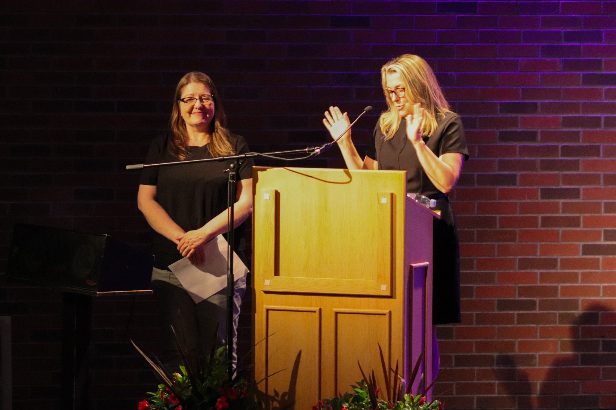 Two women standing on stage behind a wooden podium, one speaking into a microphone with hands raised, the other smiling and holding papers, against a dark brick wall background.