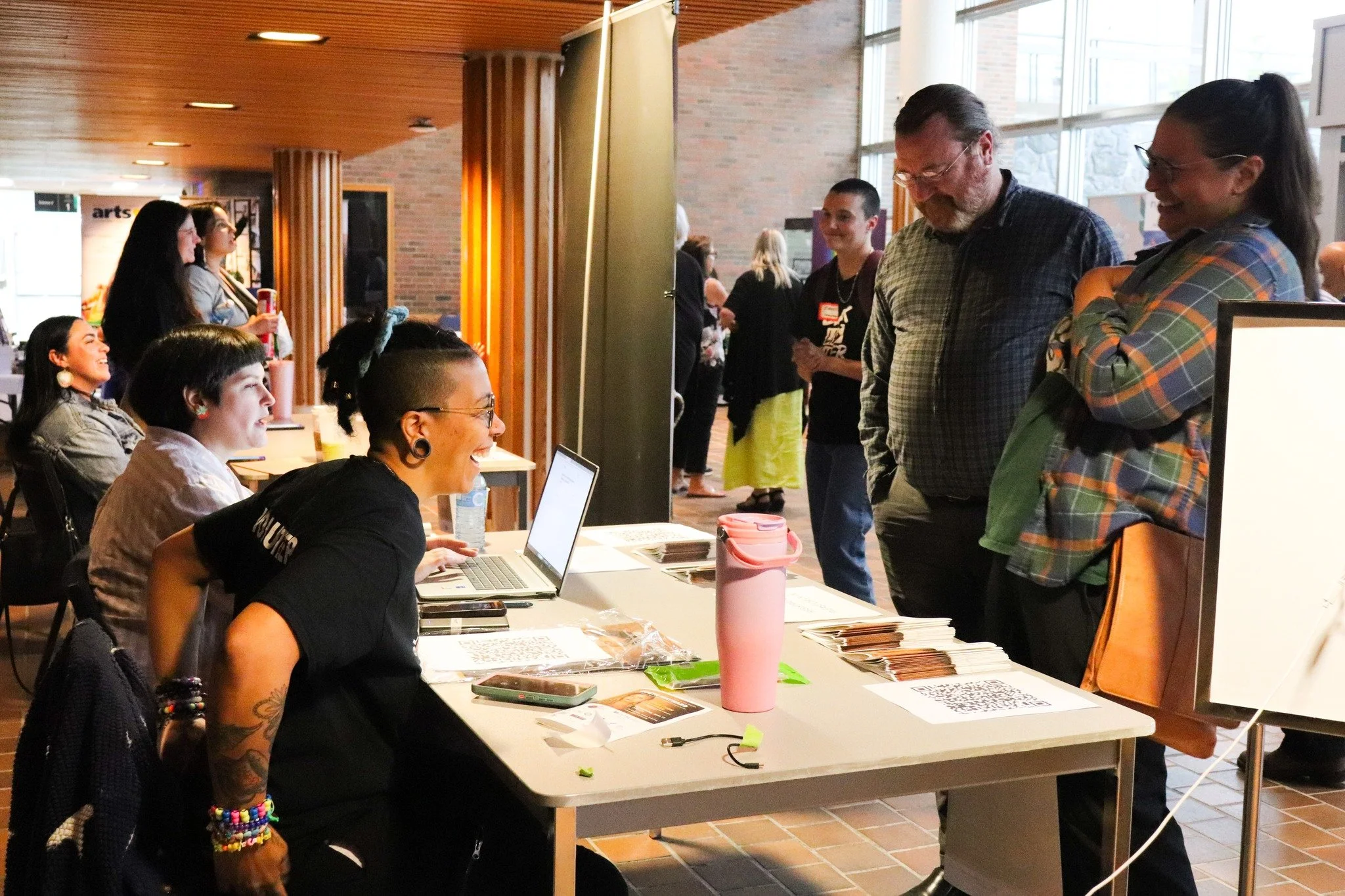People standing in line and sitting at a registration or information desk at an indoor event, smiling and interacting with staff.