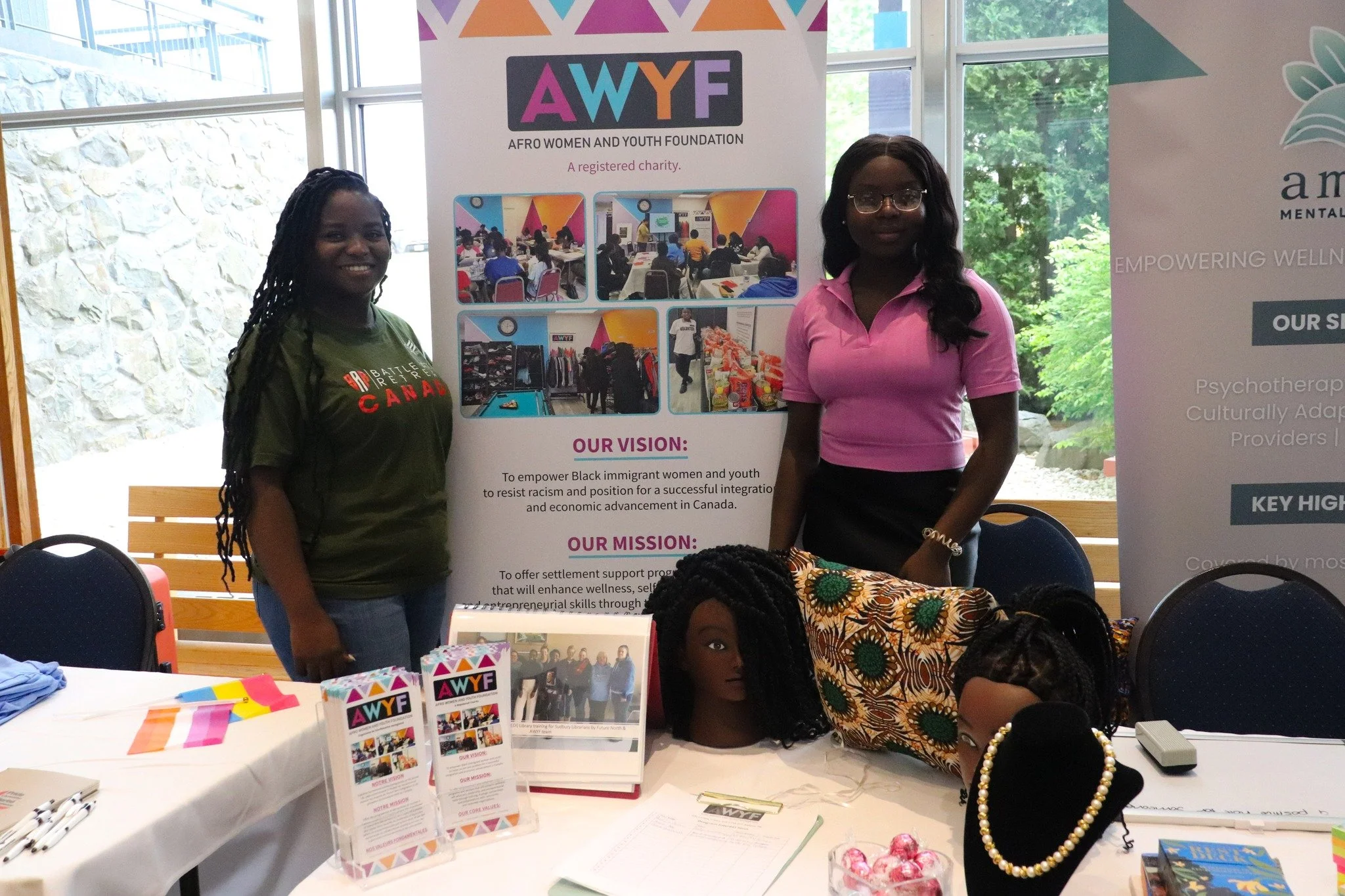 Two women stand behind a table at a community outreach booth for AFRO Women and Youth Foundation (AWYF). The table displays informational brochures, mannequin heads with wigs, and jewelry. Behind them are standing banners with AWYF branding and mission statements.