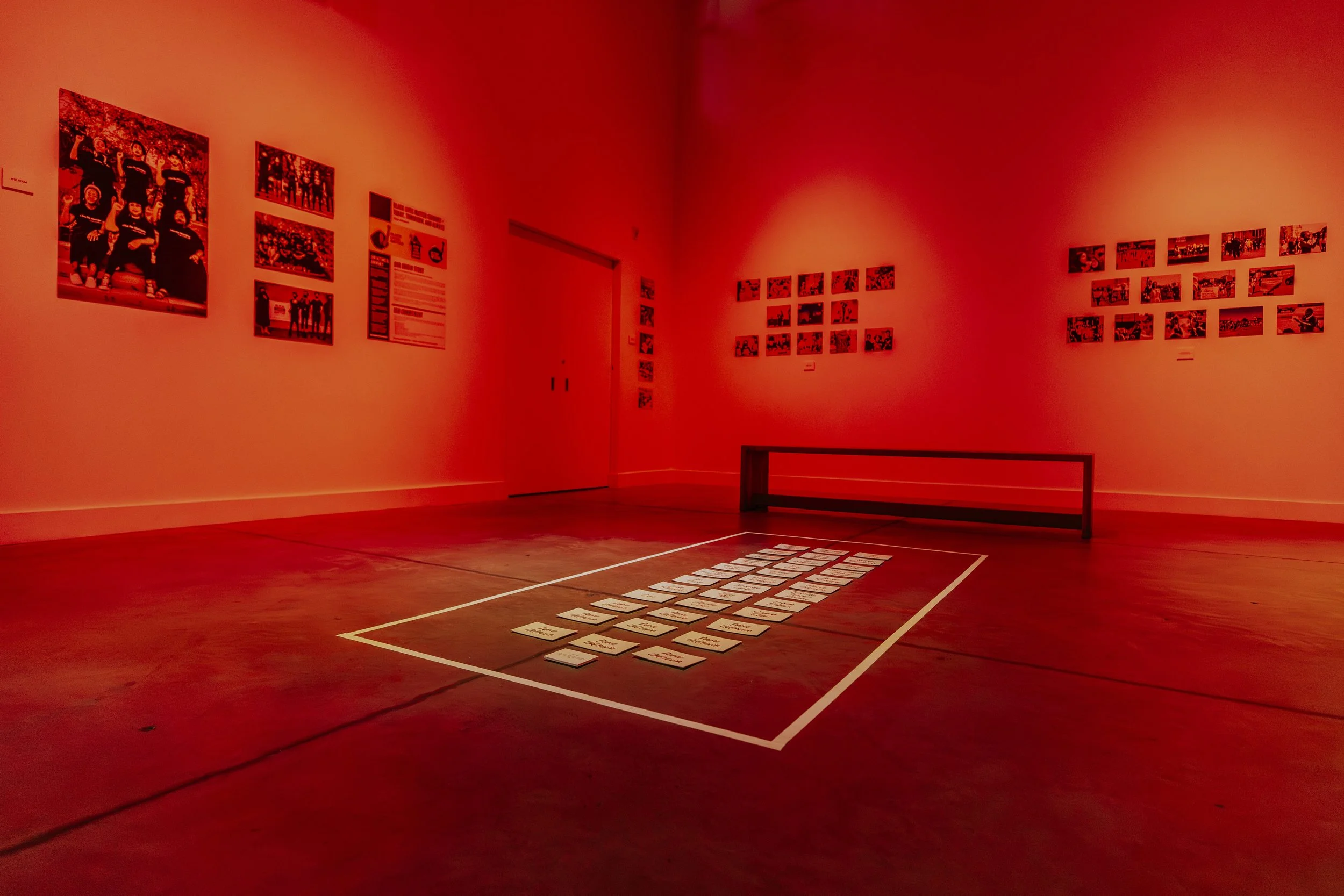 An art exhibit in a gallery space with red lighting, displaying photographs on the walls and notes laid out on the floor within a white border.