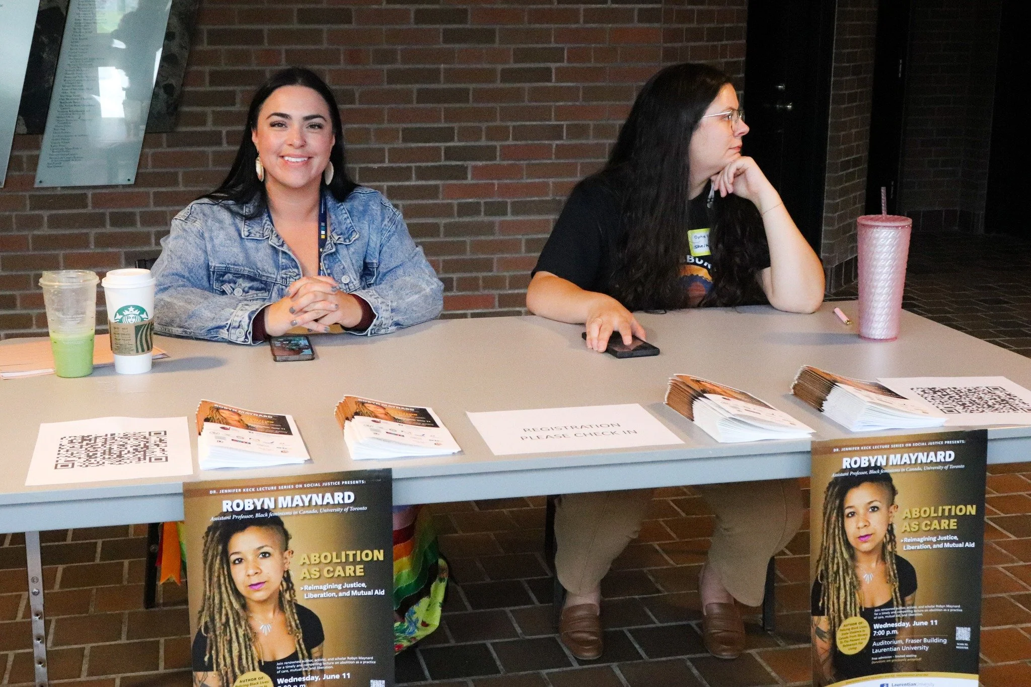 Two women sitting at a registration table with papers, pamphlets, and QR codes, set up for an event on social justice, with banners displaying information about speaker Robyn Maynard.