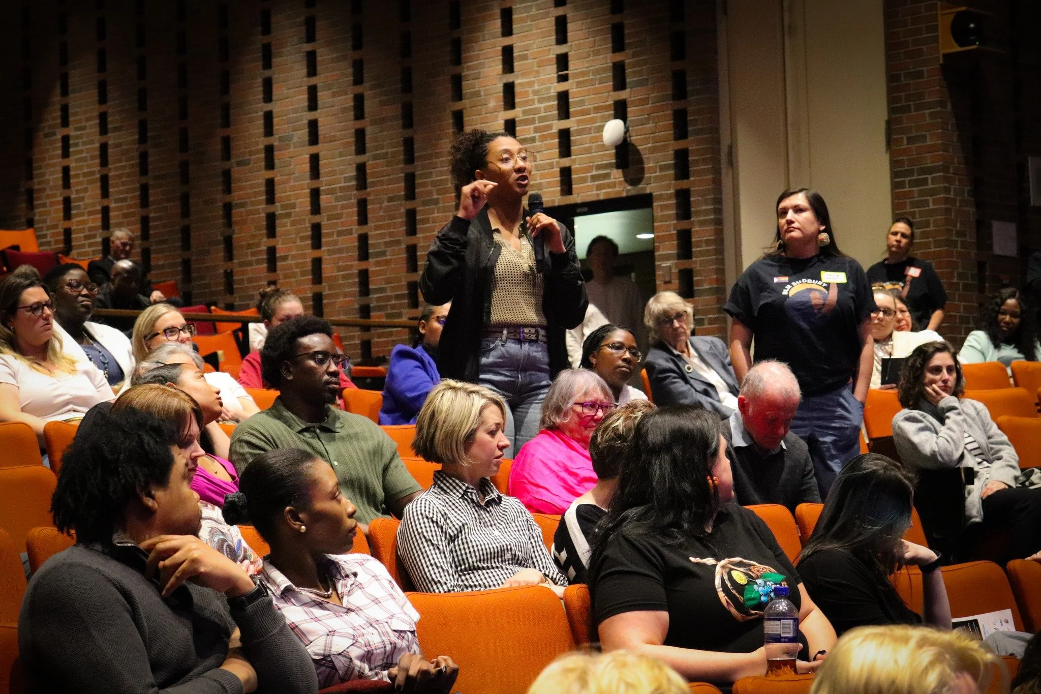A woman standing and speaking into a microphone in a crowded auditorium with brick walls, with attentive audience members seated and listening.