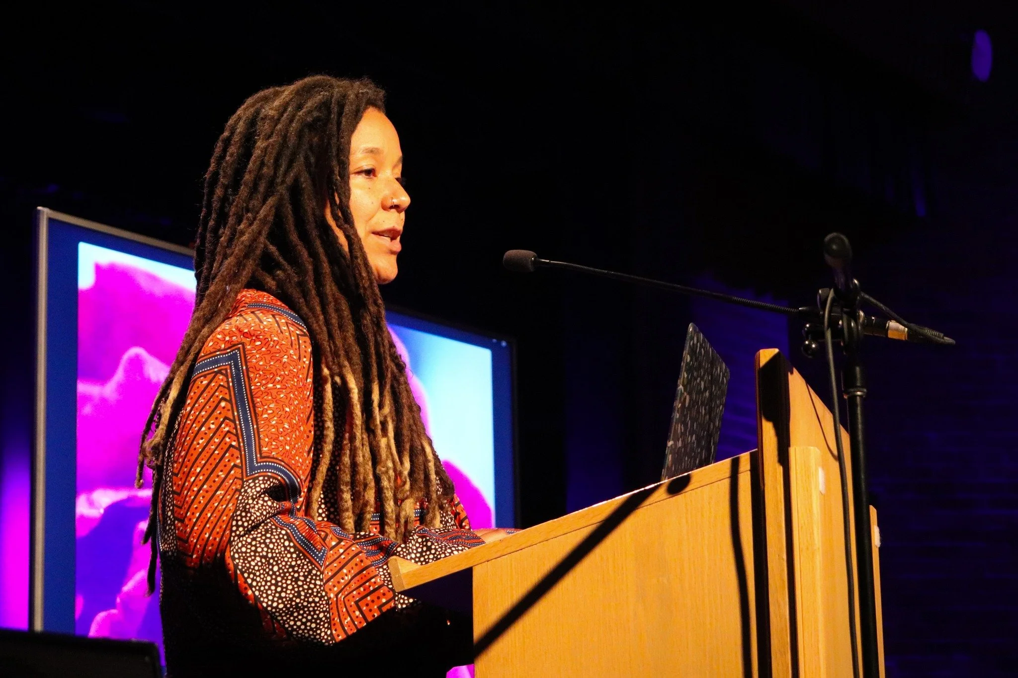 A woman with long dreadlocks stands at a wooden podium speaking into a microphone, with a screen displaying a pink and purple abstract background in the background.