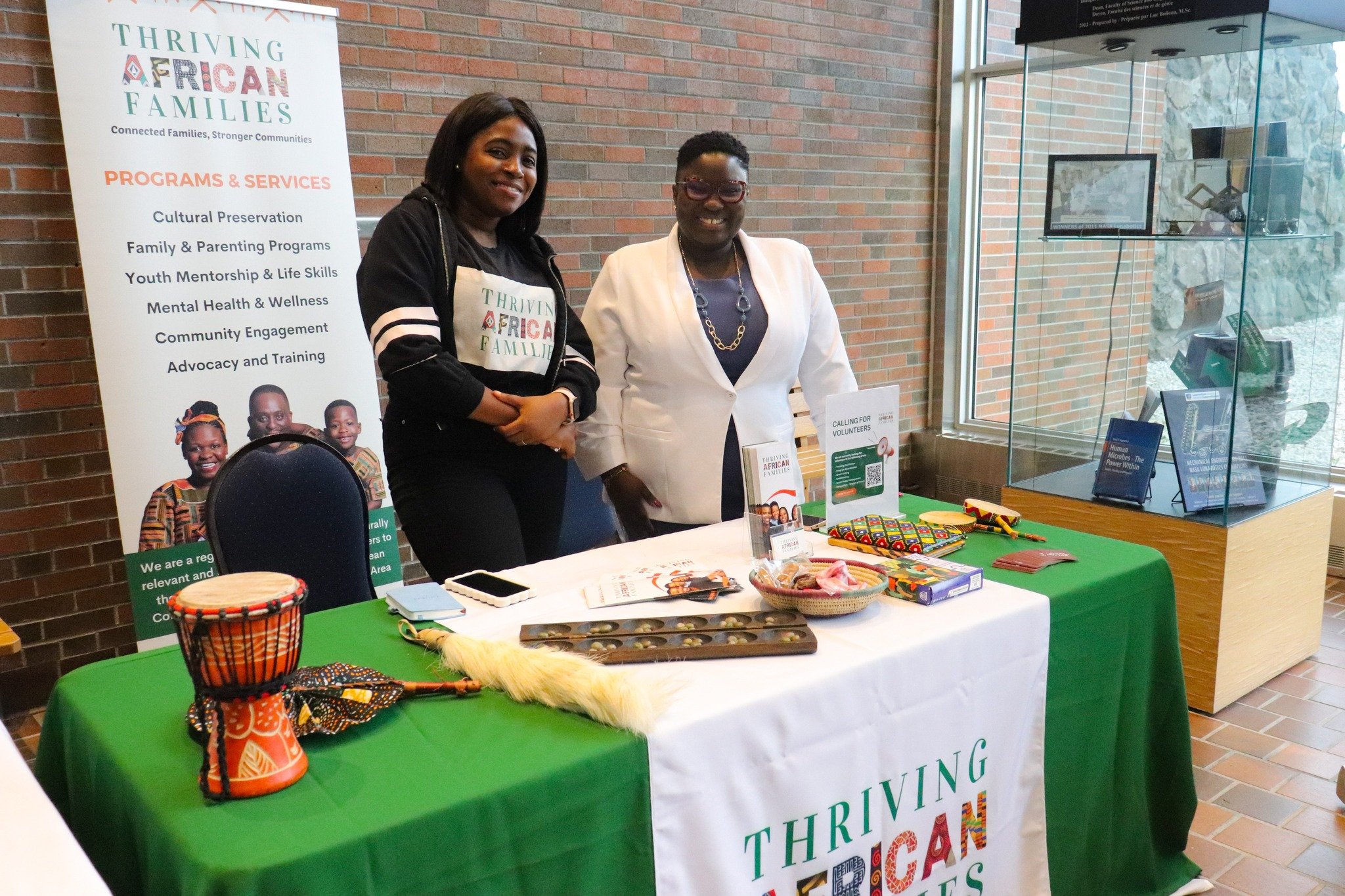 Two women stand behind a table with items related to African culture, including drums and accessories, at a booth for Thriving African Families, with a banner and display case nearby.