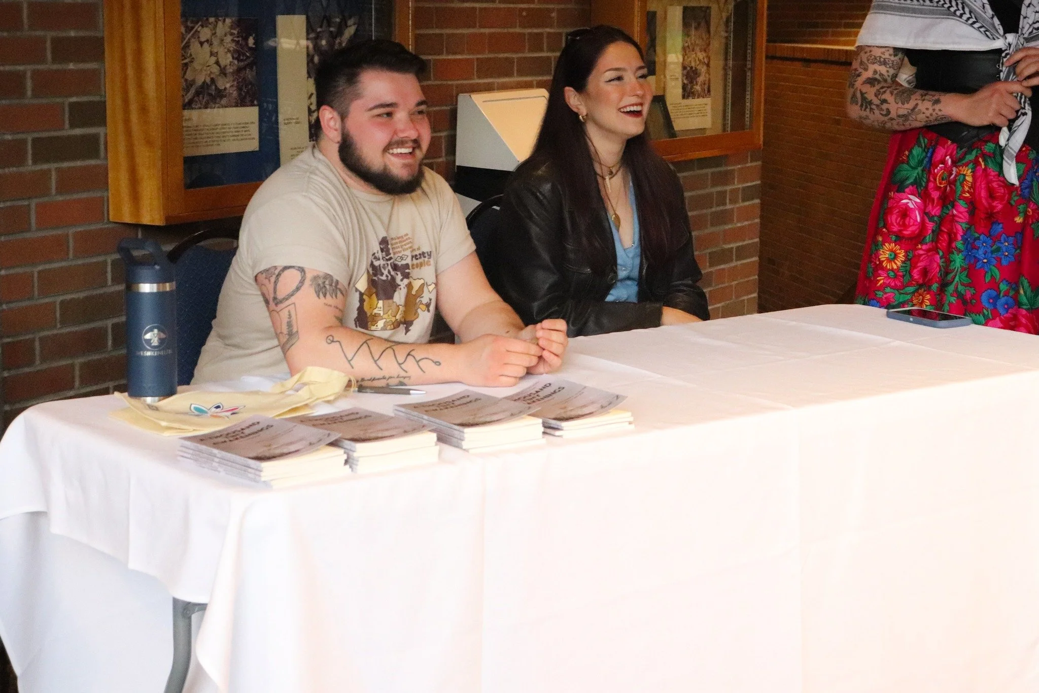 Two people sitting at a table, smiling, with pamphlets and a water bottle in front of them, in a room with brick walls.