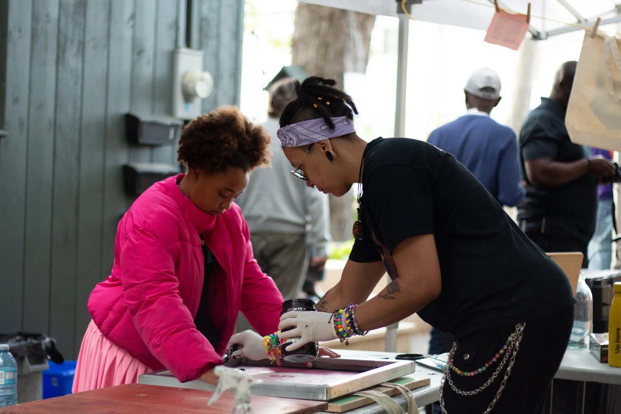 Two women working together on an art project at a crafting table, with multiple people in the background.