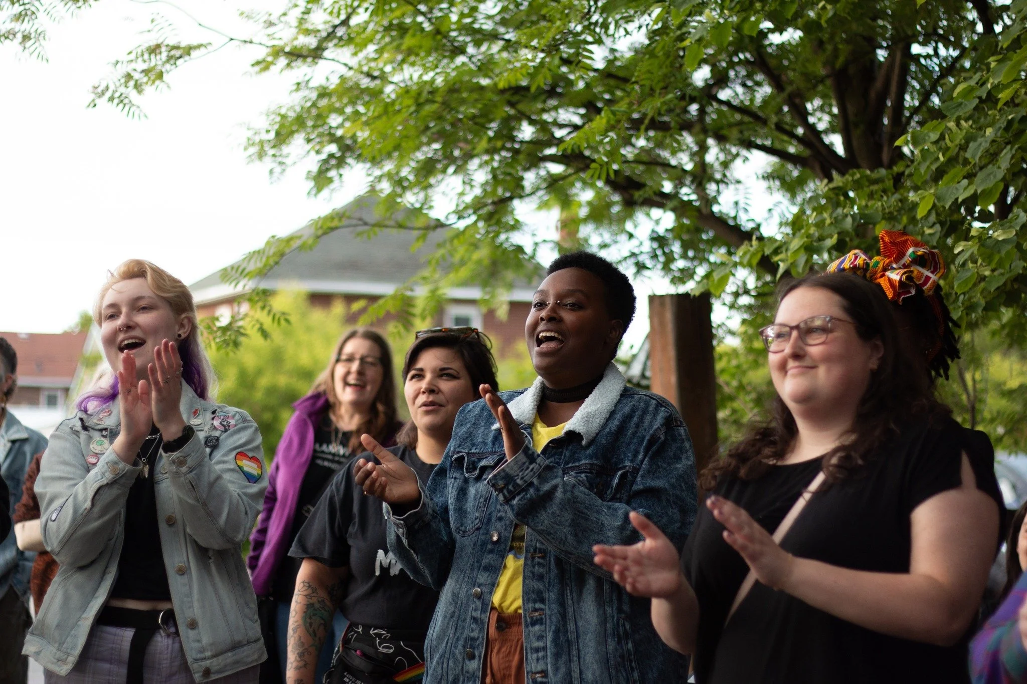 Group of diverse women outdoors, smiling and clapping, under a leafy tree, celebrating or enjoying a social gathering.