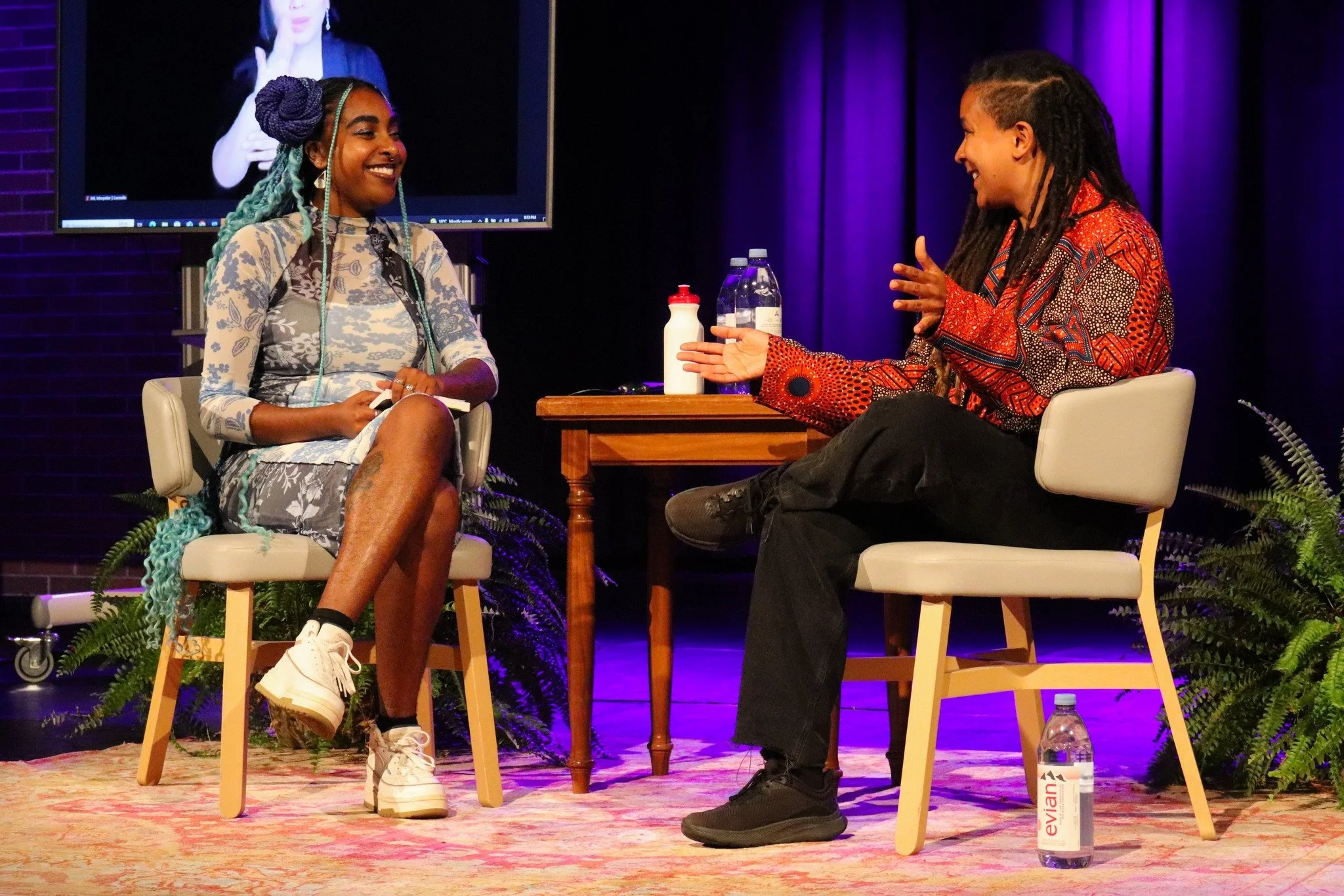 Two women having a conversation on stage, sitting in chairs with small tables between them. One woman has braided hair with teal extensions, wearing a patterned dress and white sneakers, smiling. The other woman has dreadlocks, wearing a colorful patterned shirt, gesturing with her hands. The stage has purple lighting, with a water bottle on the floor and plants nearby.