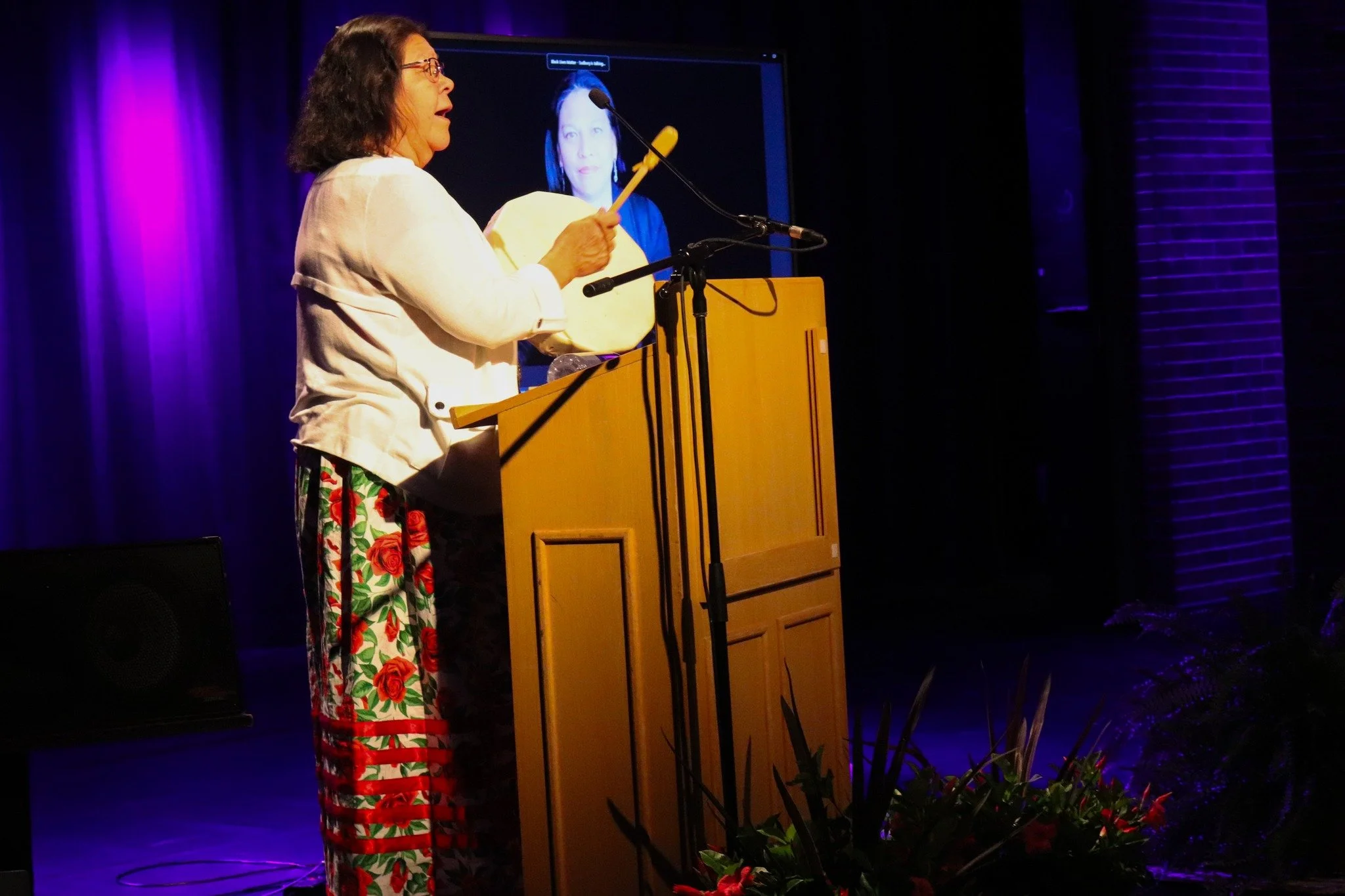 A woman with glasses and dark hair, wearing a white jacket and floral pants, stands at a wooden podium on a stage. She is playing a tambourine with one hand, while speaking into a microphone. Behind her, a large screen displays her image and a purple curtain is visible in the background. Stage plants are at the front of the stage.