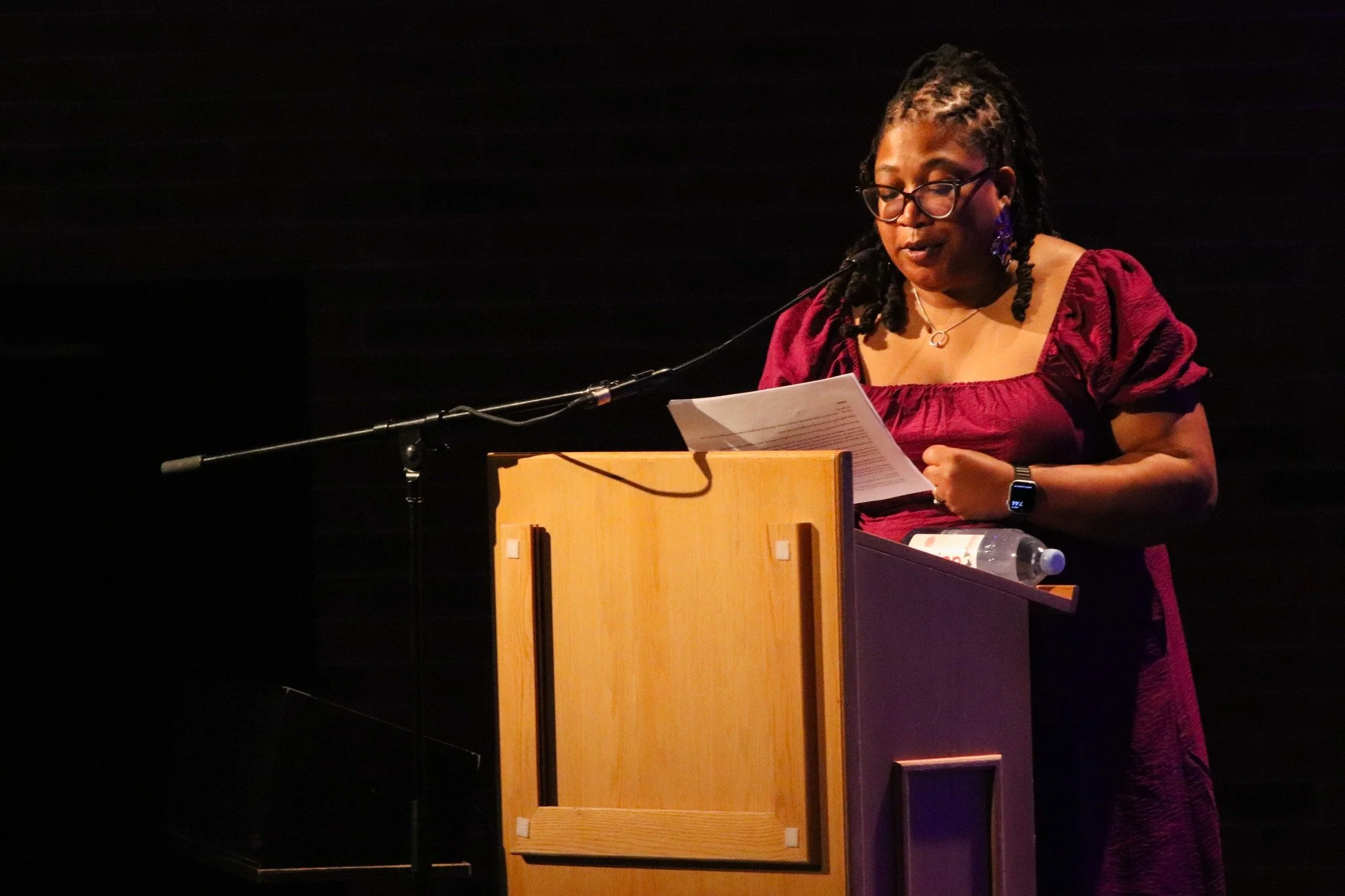 A woman with glasses and dreadlocks, wearing a burgundy dress, standing at a podium with a microphone, reading from papers with a water bottle nearby, against a dark background.