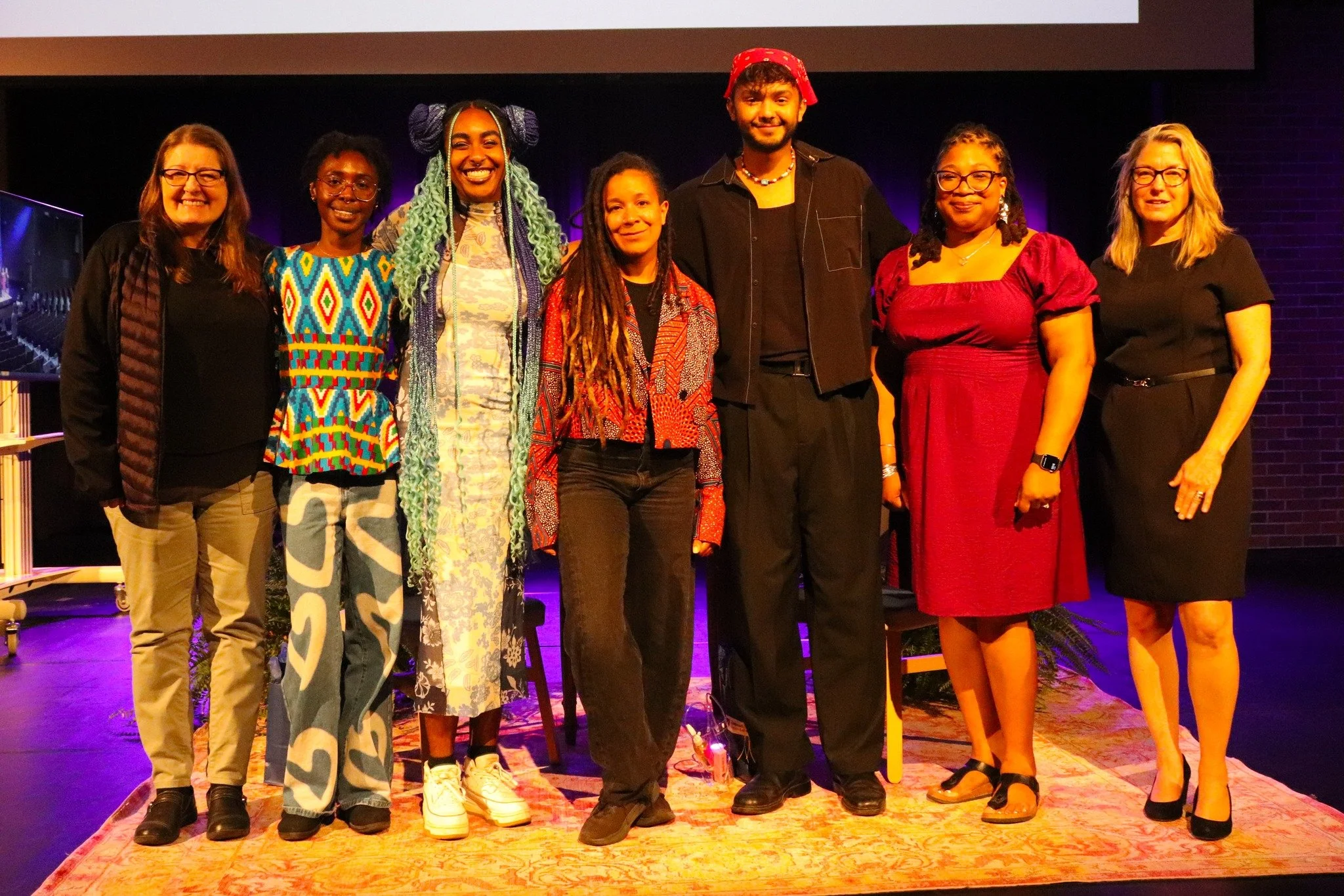 Group of eight diverse women standing on a colorful rug on stage, smiling for a photo, with purple and dark background lighting.