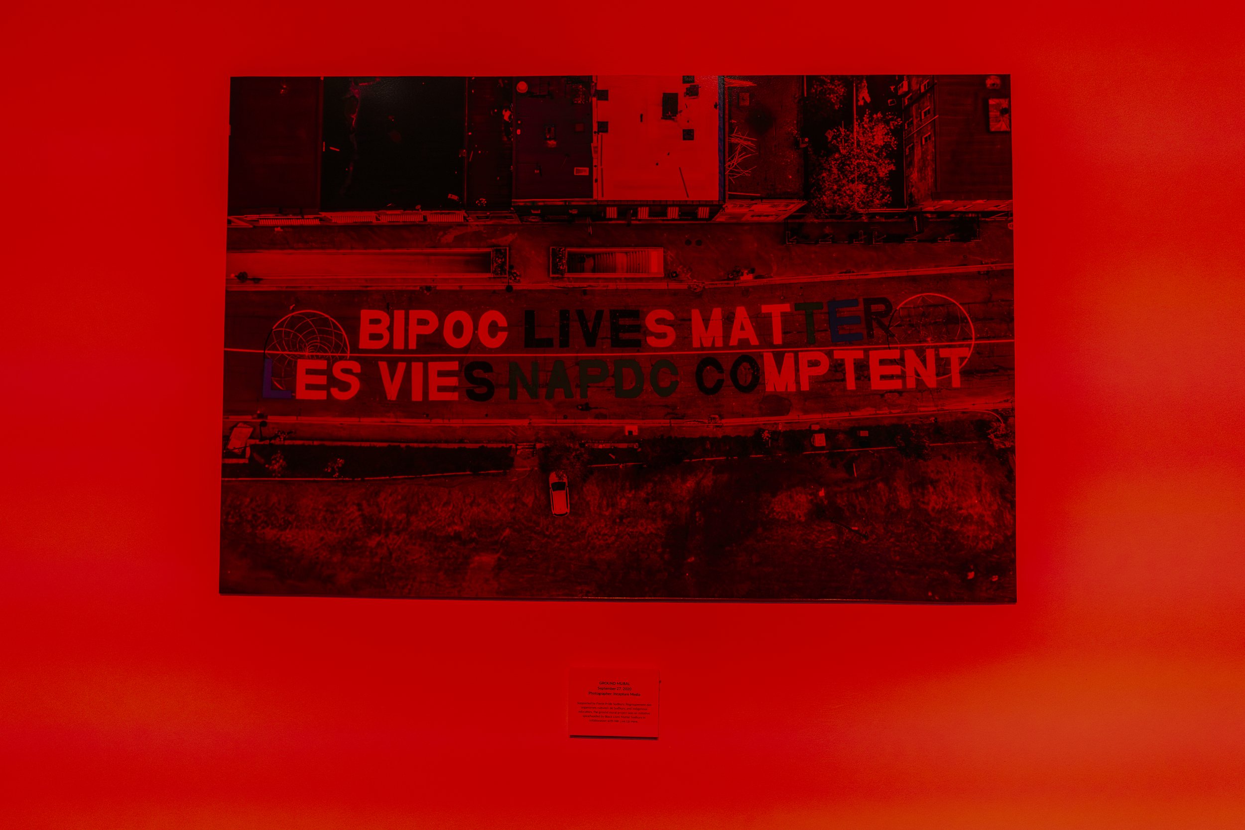Aerial view of a city street with large mural that reads 'BIPOC LIVES MATTER ES VIES NAPDC COMPTENT' in bold red and black letters.