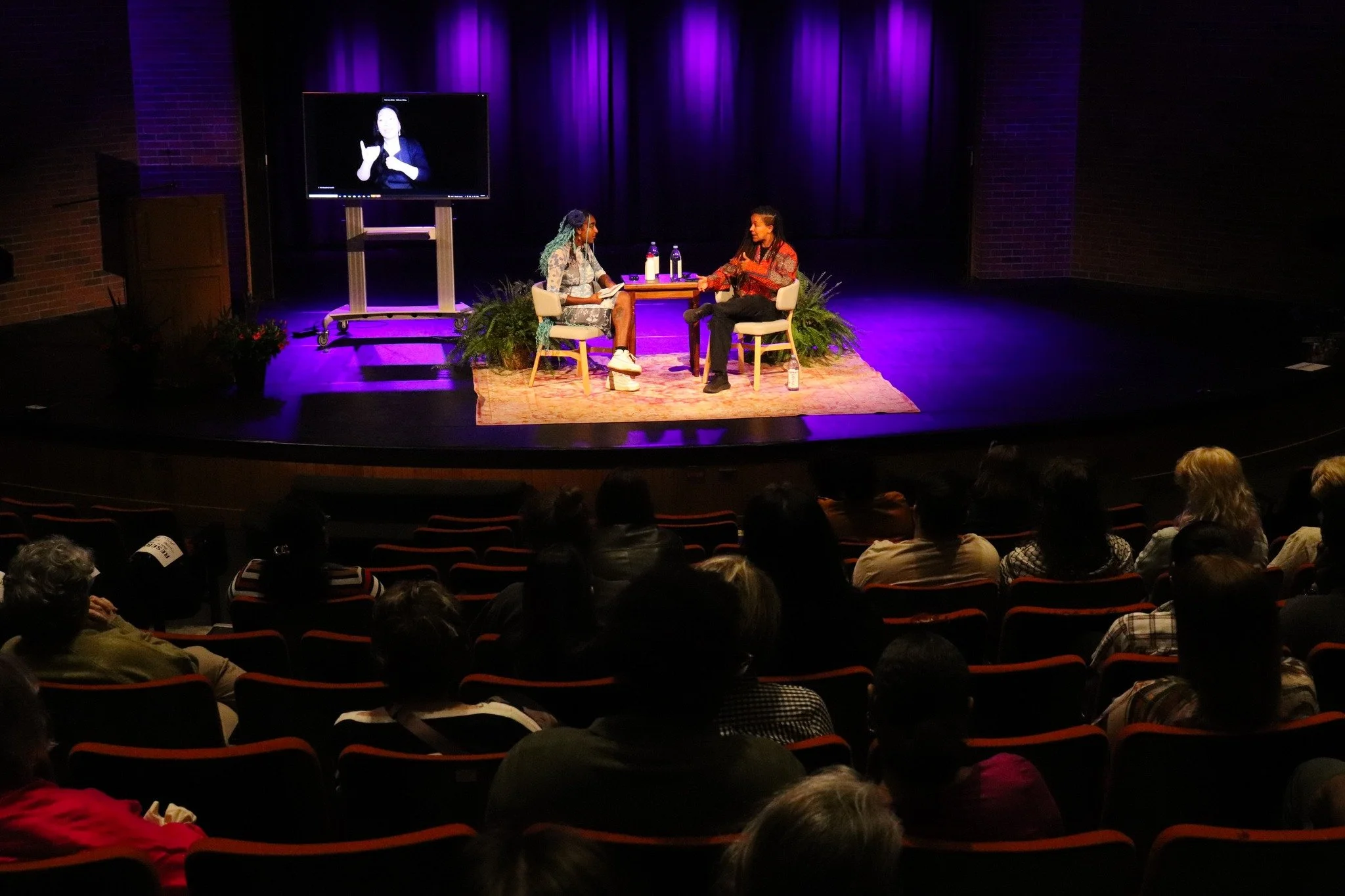 Two women sitting and talking on stage during a presentation or interview, with a small table, water bottles, and a large screen behind them showing sign language interpretation, in an auditorium with an audience.