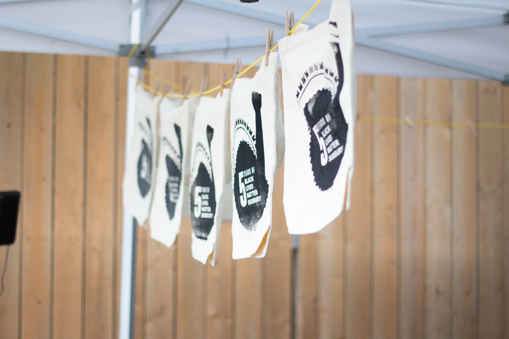 Facsimile artwork or banners hanging on a clothesline inside a structure with a wooden wall, featuring bold black and white graphic logos and text for an event related to Black Lives Matter.