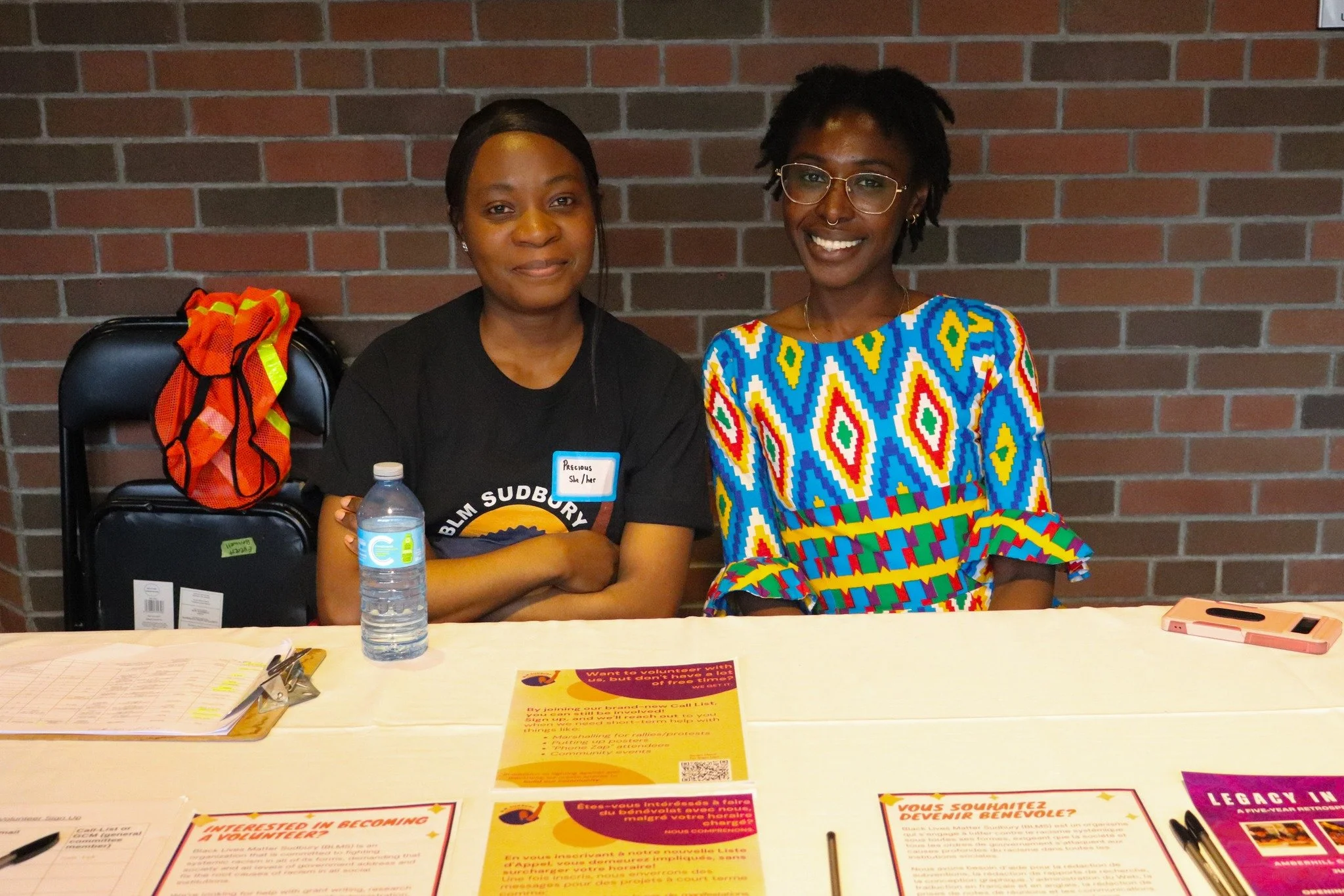 Two women sitting at a table with brochures and a water bottle, against a brick wall background. One woman is wearing a black t-shirt with a badge, and the other is in a colorful patterned dress.