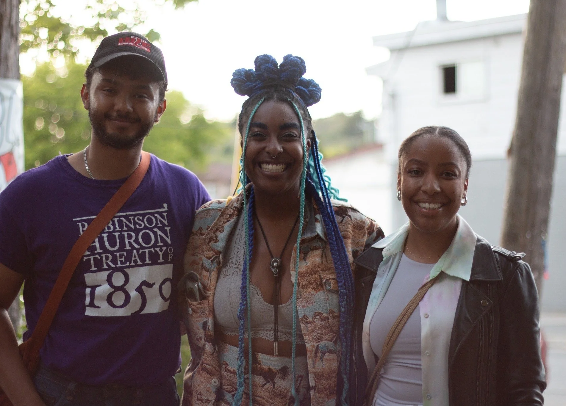 Three friends standing together outdoors, smiling at the camera. Behind them are trees and a white house.
