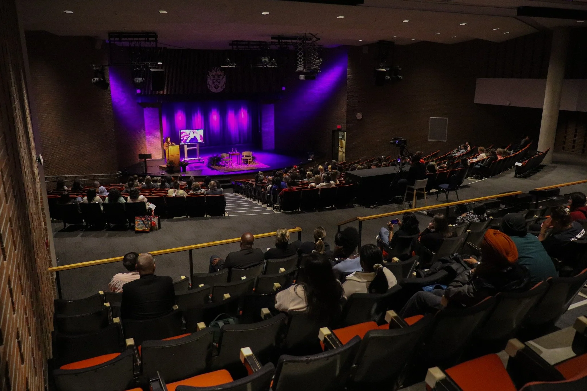 Audience seated in an auditorium watching a presentation on stage with purple lighting and a screen.