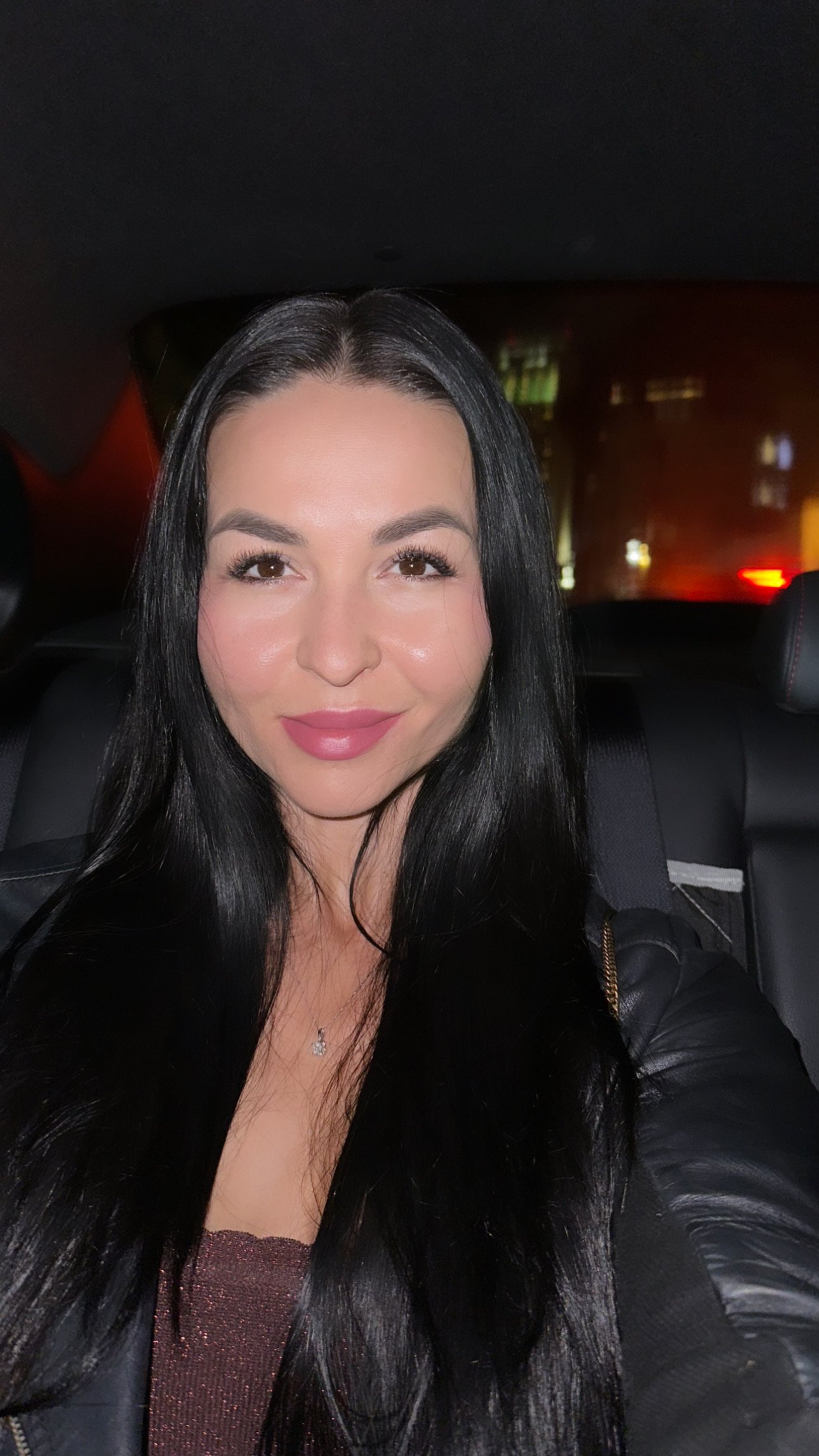 A woman with long black hair taking a selfie inside a car at night, with city lights visible through the window behind her.