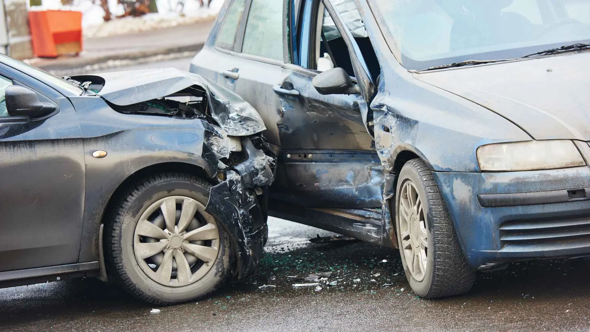 Two cars involved in a collision, showing front-end damage to the black car and side damage to the blue car, on a wet road.