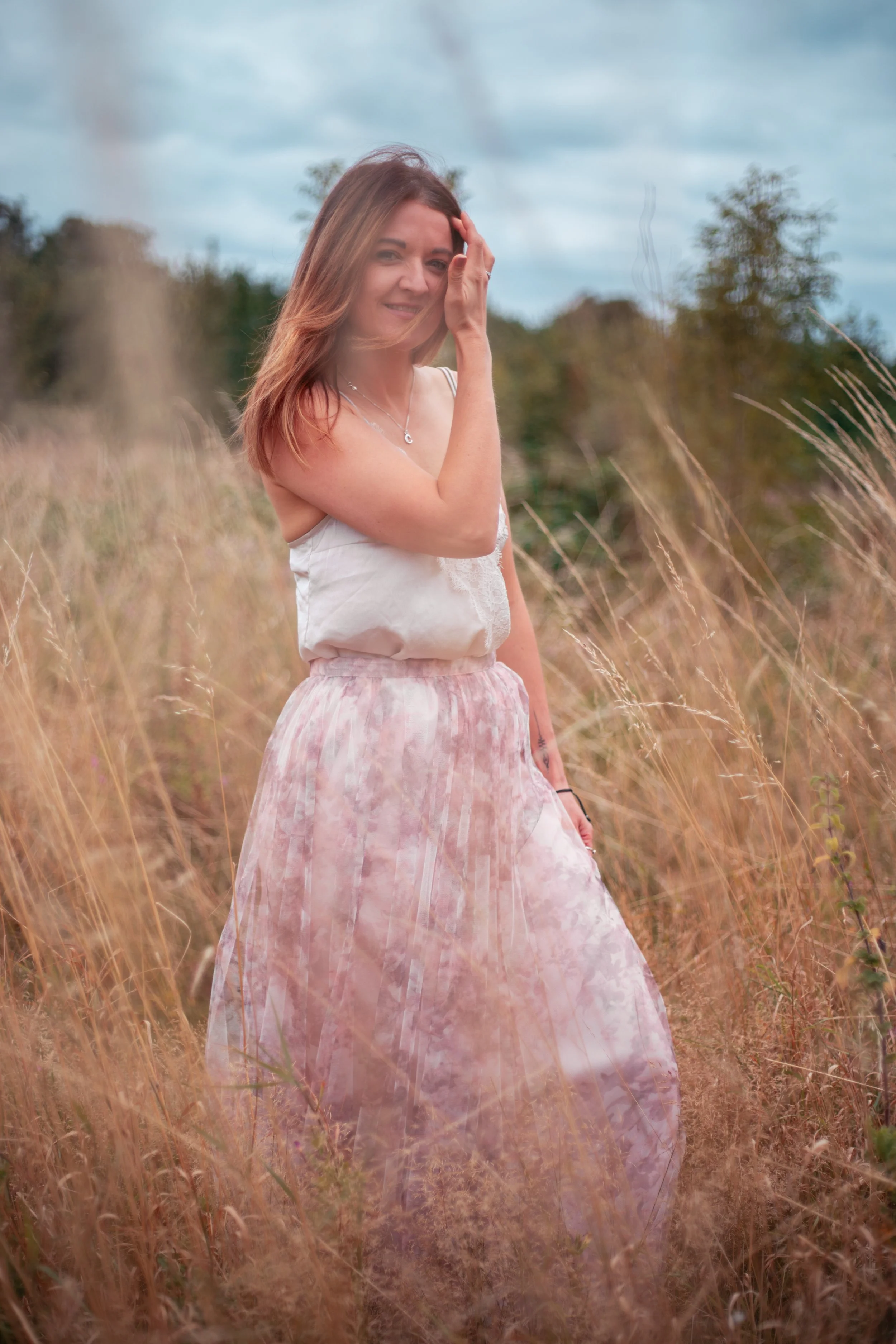 A woman standing in a field of tall, golden grass, smiling and touching her hair, wearing a white sleeveless top and a long pink and white skirt with a cloudy pattern. Natural beautiful portrait.