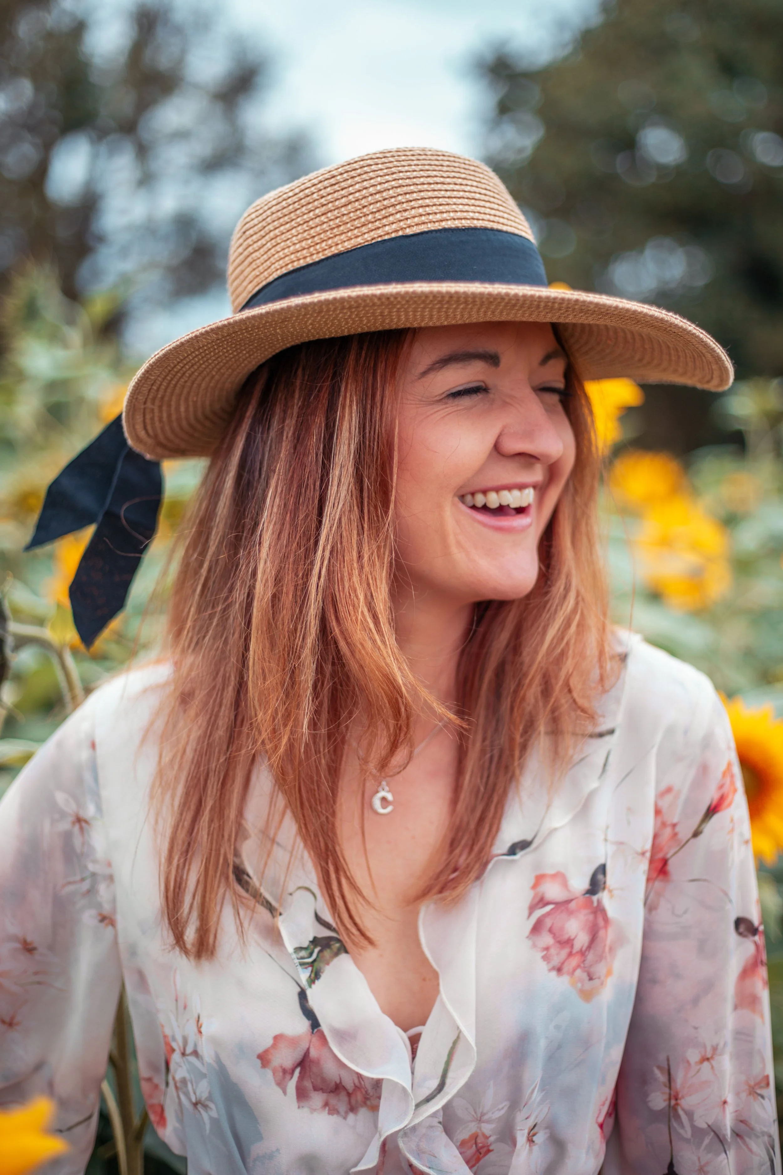 A woman with red hair wearing a straw hat with a black ribbon, smiling in a sunflower field. Natural beautiful portrait.